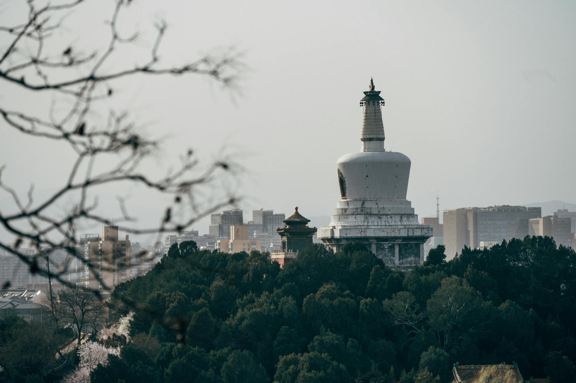 Overcast view of the White Pagoda in Beihai Park, Beijing, amid urban skyline and lush greenery.