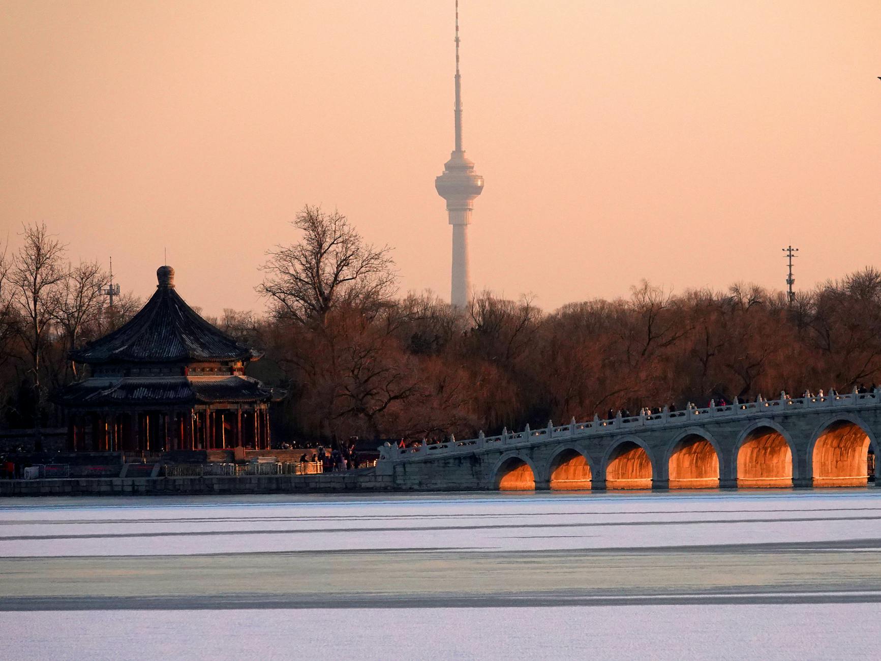 Orange hues glow on the 17-Arch Bridge and pagoda at Kunming Lake, Beijing at sunset, with the CCTV Tower in the background.