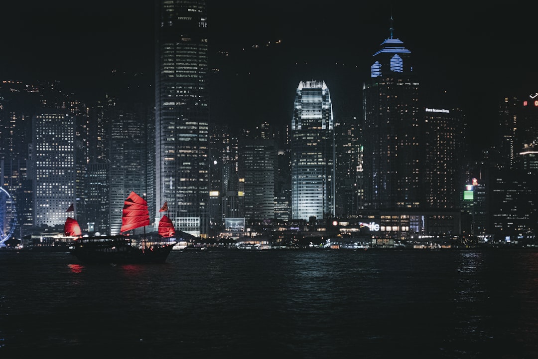 Red-sailed junks float on dark water near city lights.