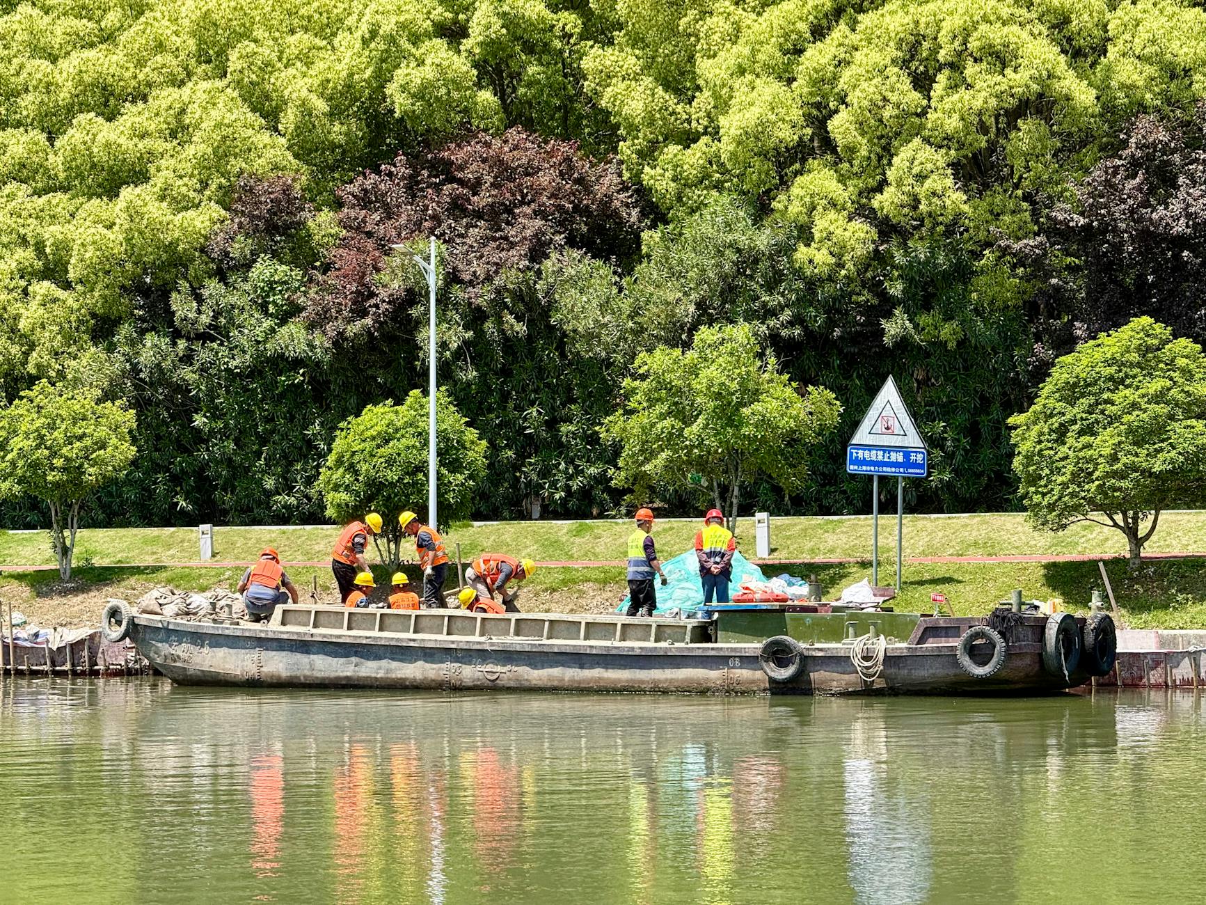 Workers with safety vests and helmets operating on a boat docked by lush greenery in a park.
