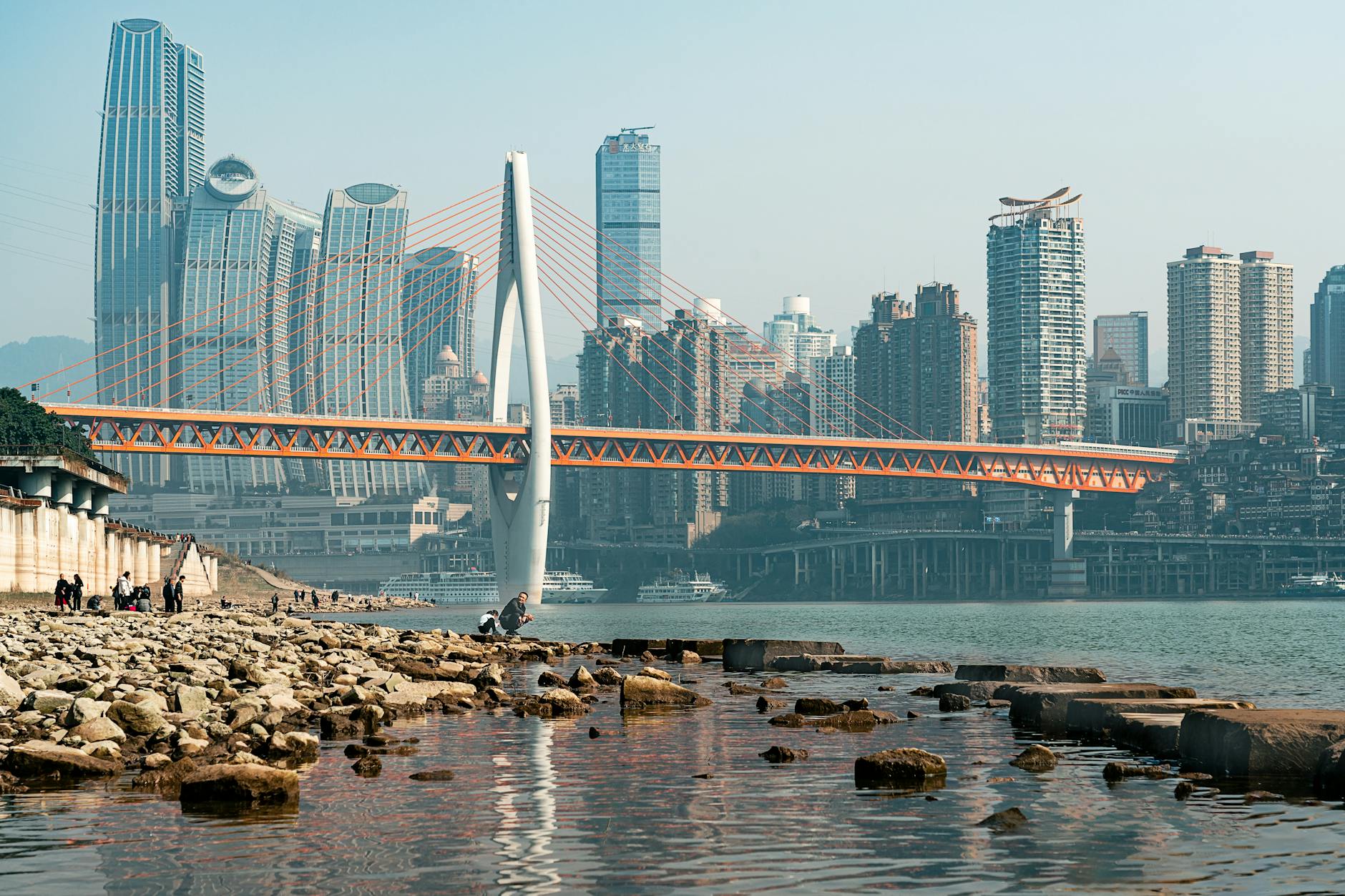 Chongqing skyline featuring the Qiansimen Bridge over a calm river with modern architecture.