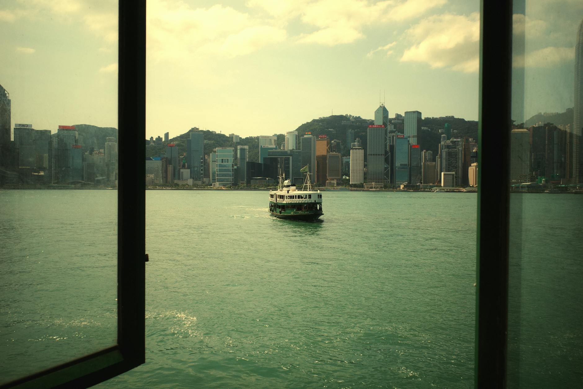 View through a window of a ferry boat approaching Hong Kong city skyline on a sunny day.
