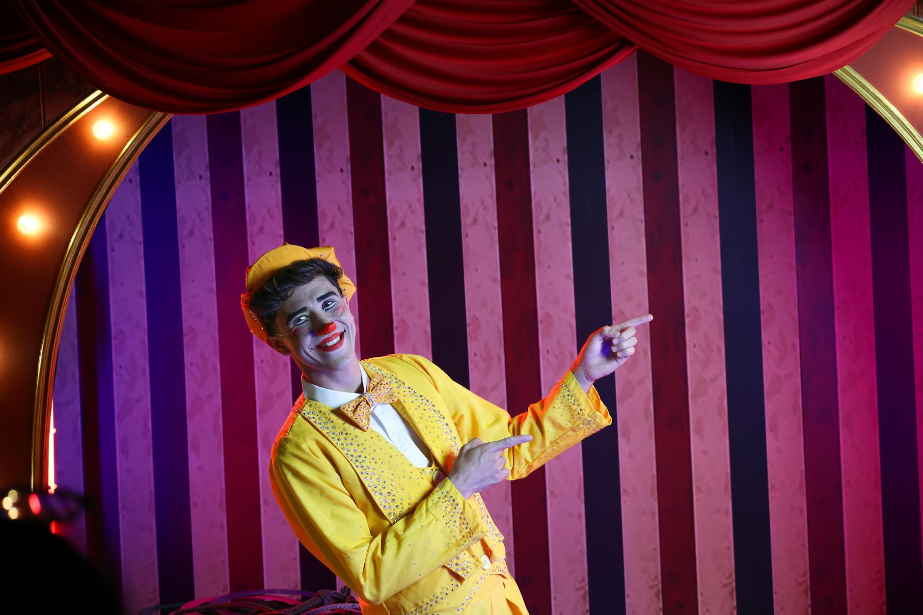 Cheerful clown in a vibrant yellow suit pointing during a stage performance, against a striped background.