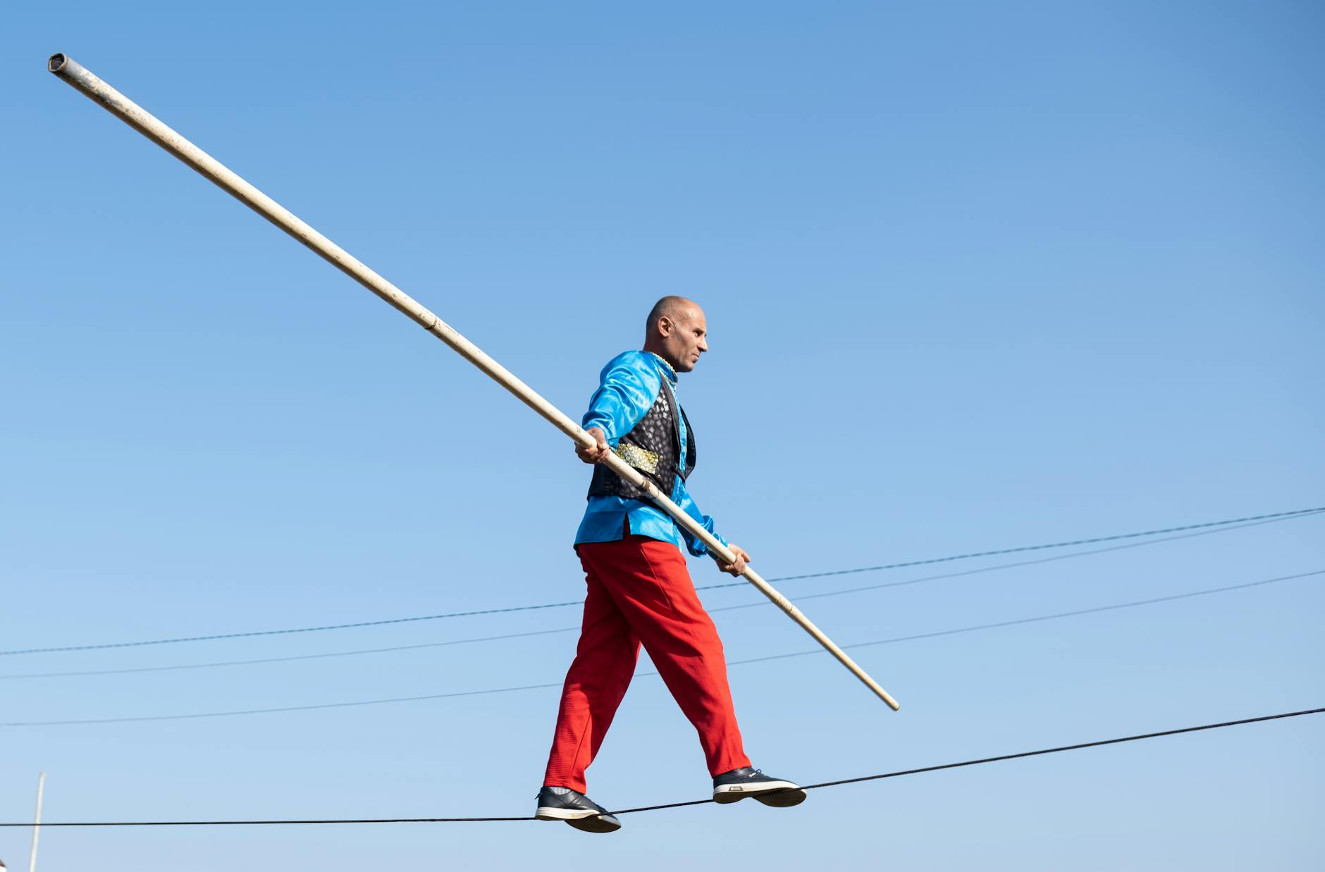 Circus performer walking on a tightrope with a balancing pole against blue sky.