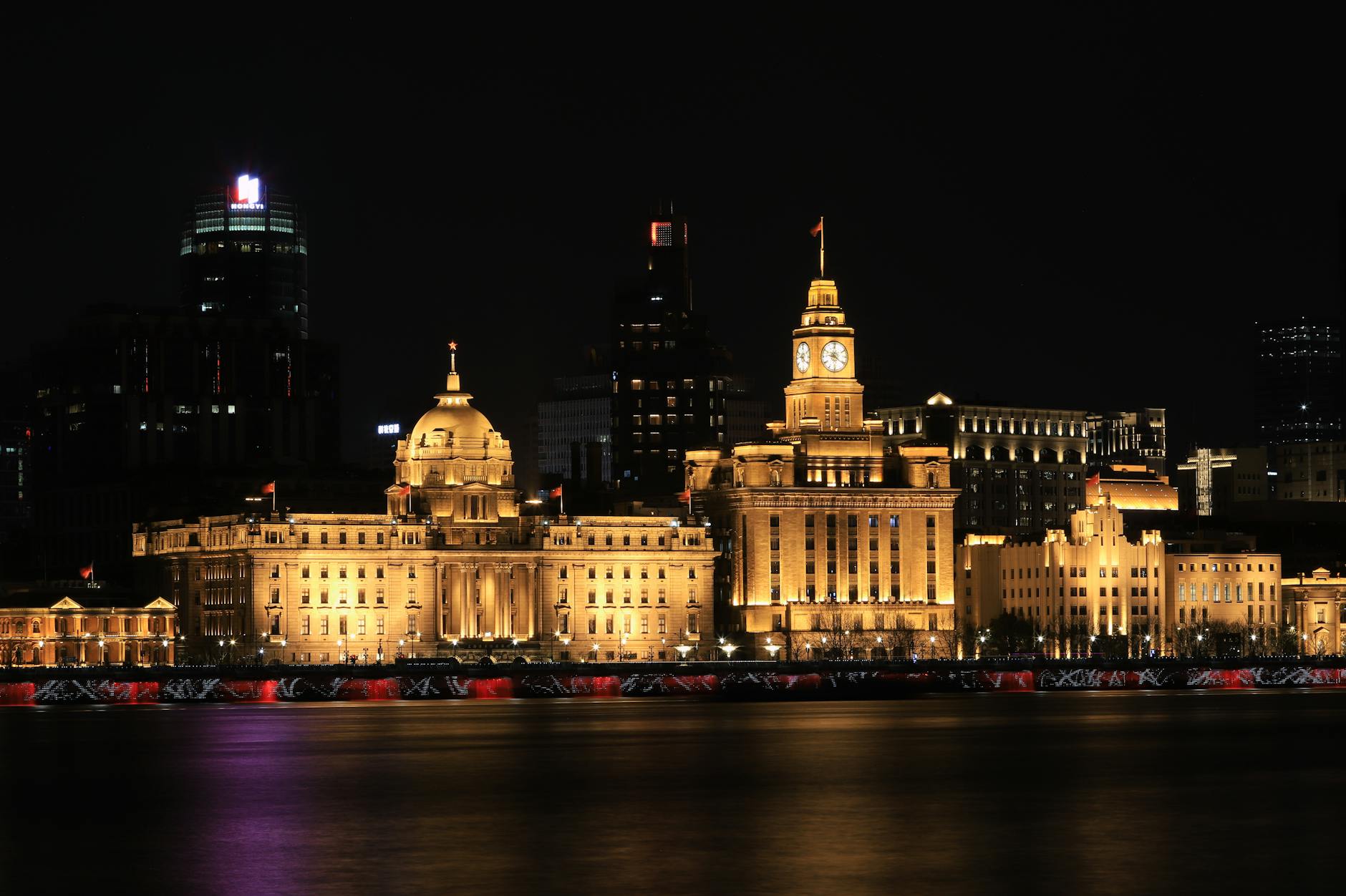 Illuminated historic buildings along The Bund in Shanghai at night, reflecting over water.