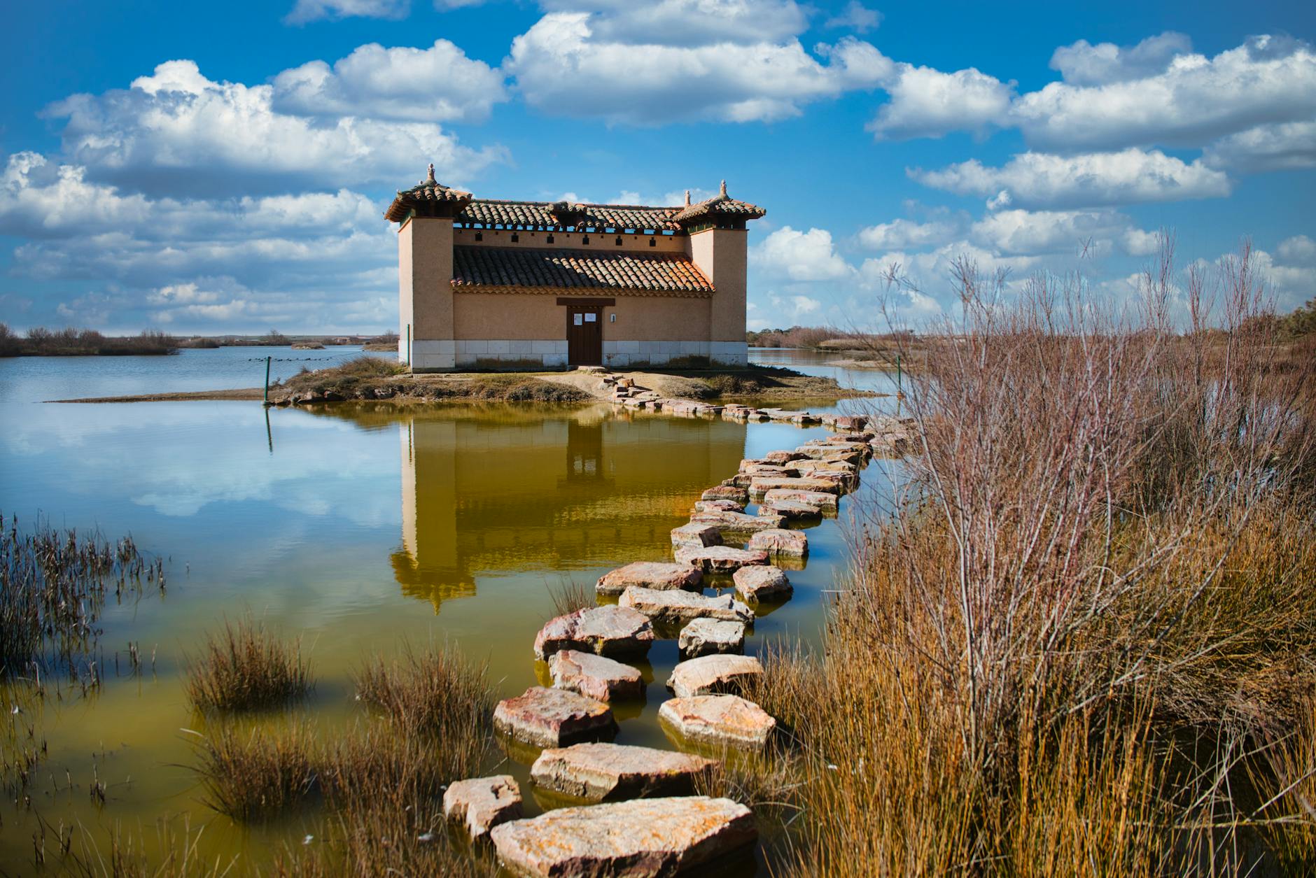 Picturesque traditional building by a serene lake in Villafáfila, Spain, featuring a stone path under a blue sky.