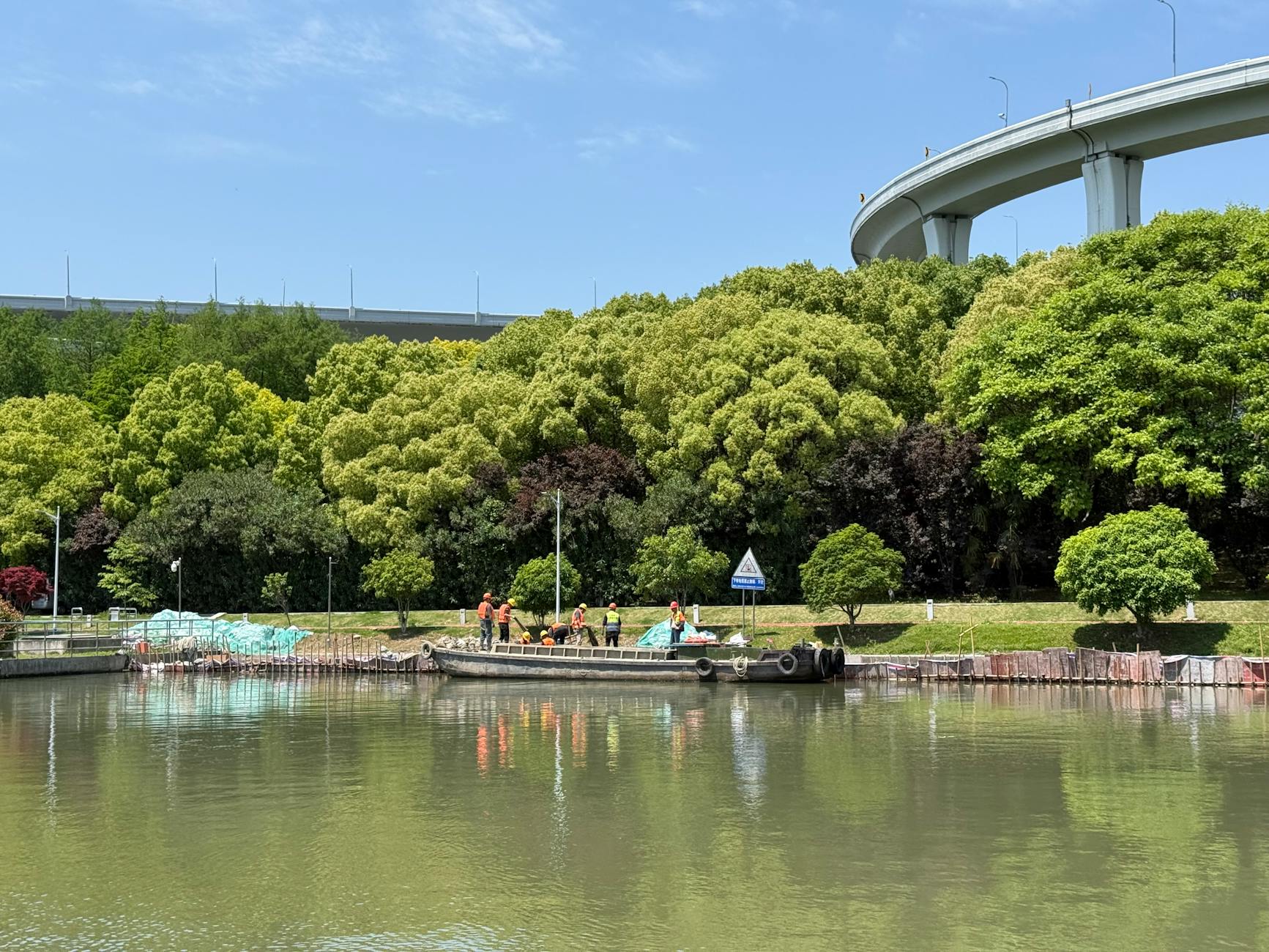 Workers on a boat in a scenic canal under a highway bridge with lush greenery.