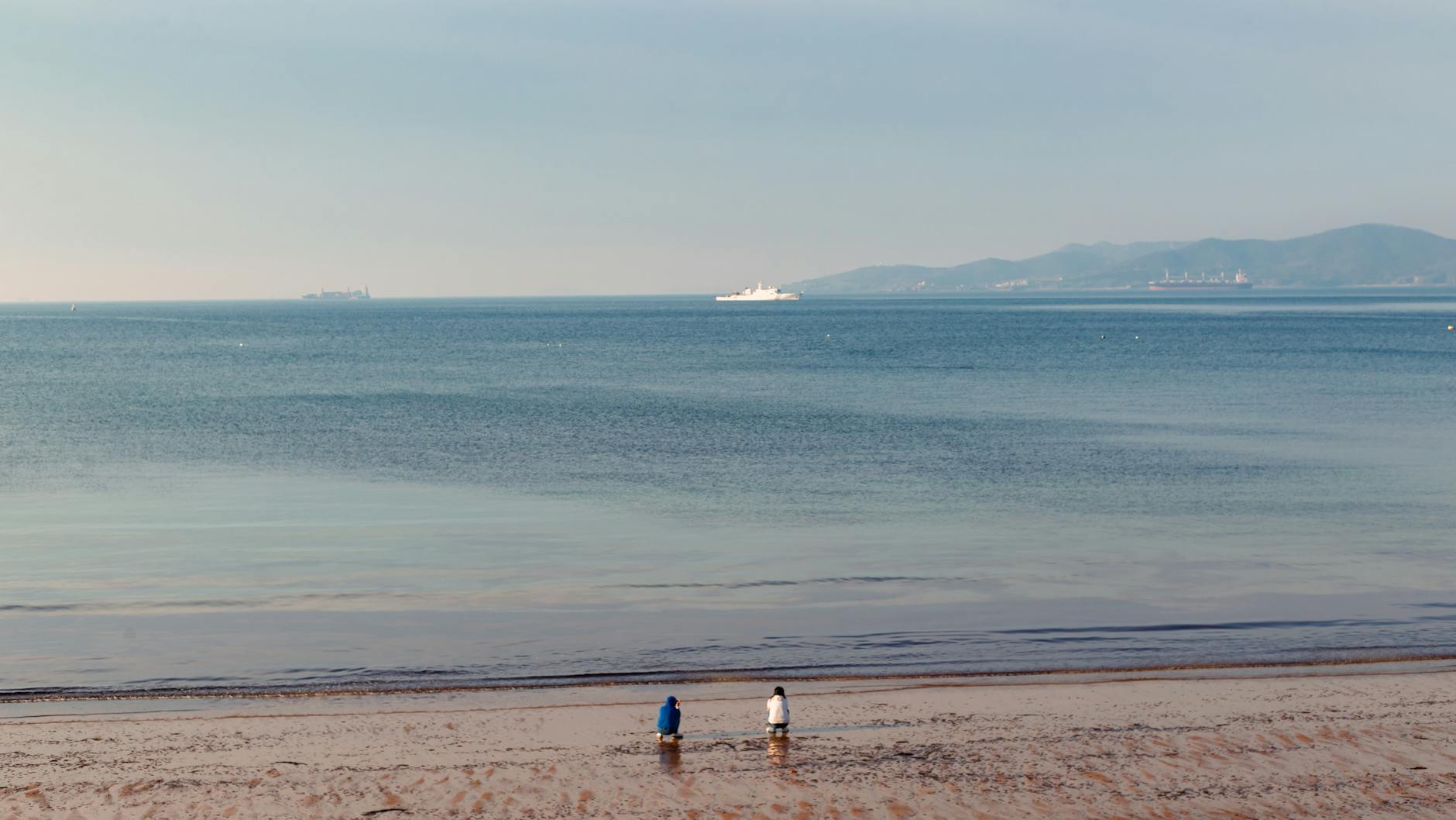 Serene beach scene with two people enjoying the view, distant ships, and a clear horizon.