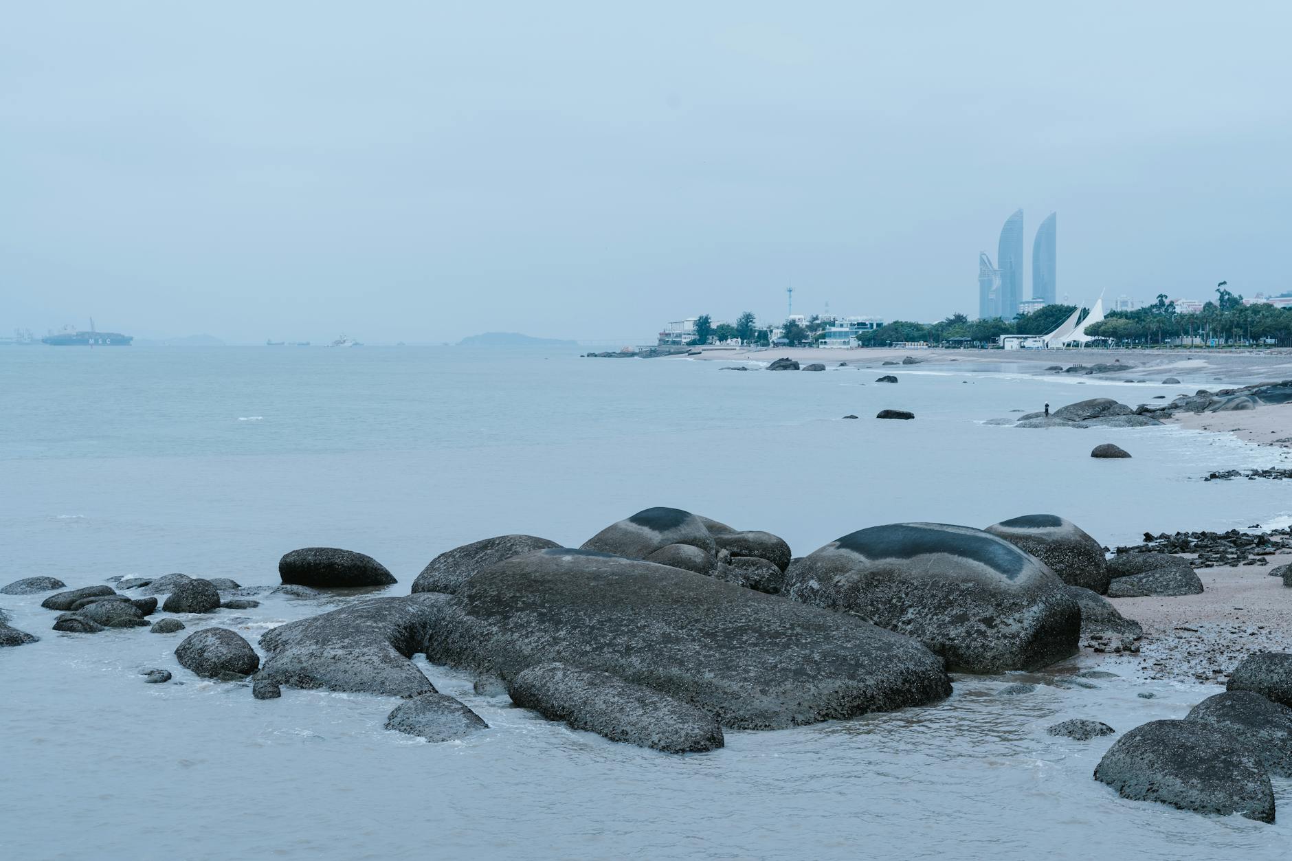 Rocky shoreline with cityscape in the background and calm ocean waves.
