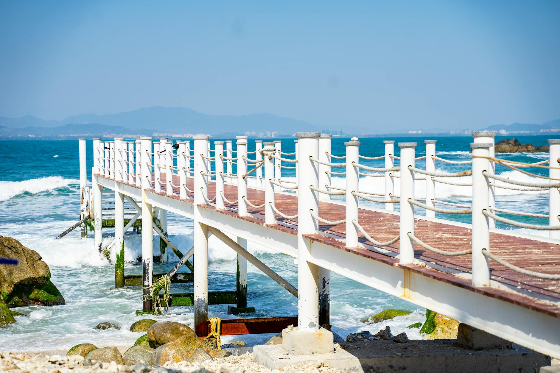 A wooden pier extends into the ocean with stunning coastal and mountain views.