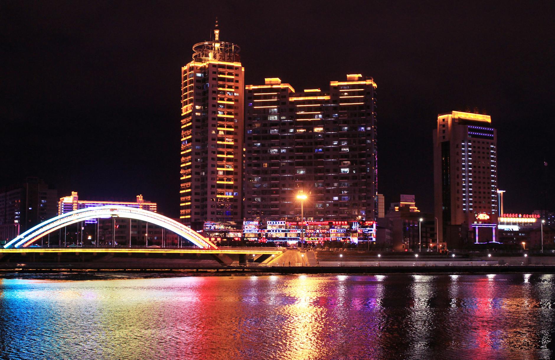 A lively cityscape featuring a bright, illuminated bridge reflected in a river at night.