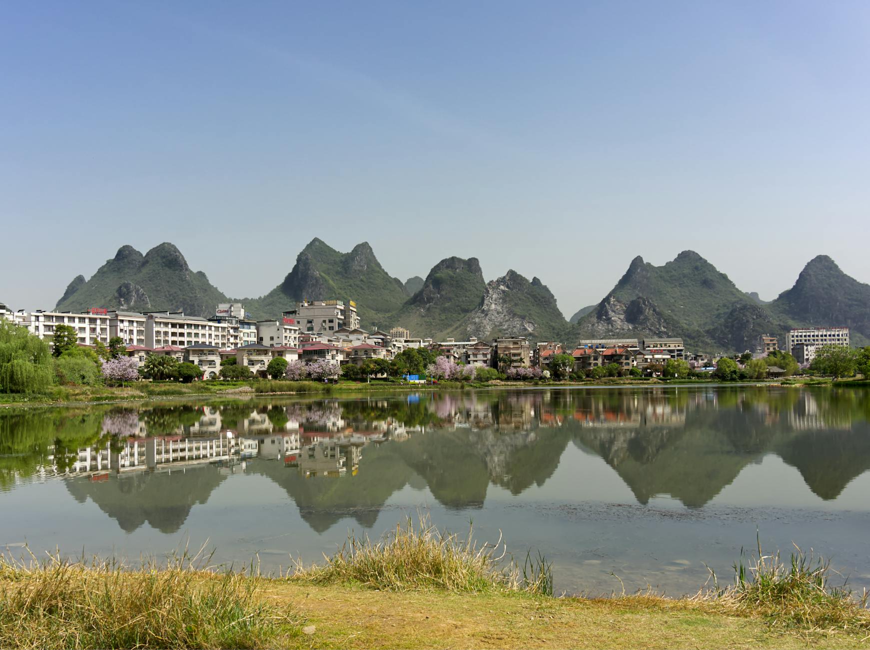 Scenic view of karst hills reflecting in a lake surrounded by buildings and greenery.