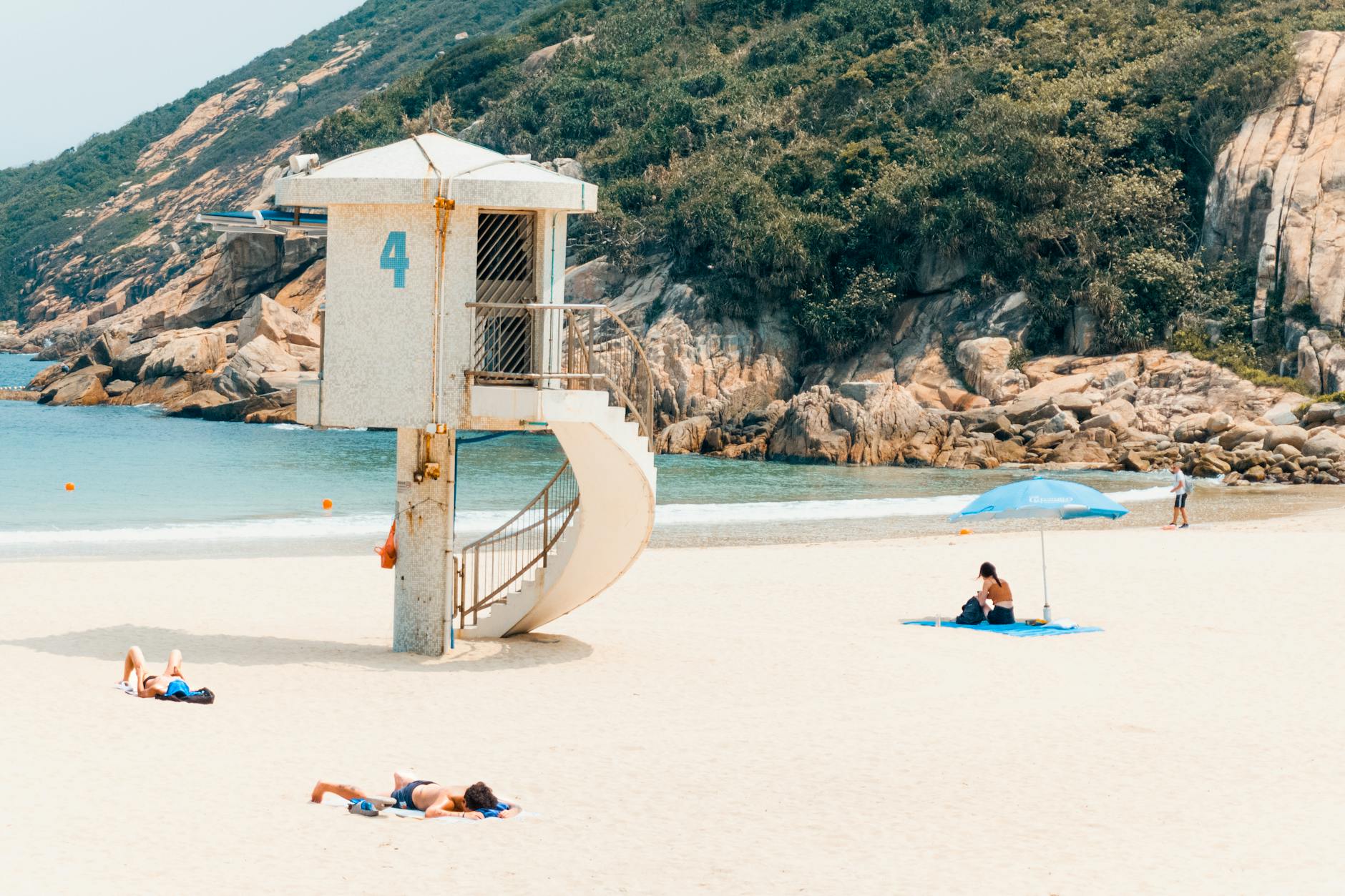 Sunlit view of Shek O Beach featuring a lifeguard hut, sunbathers, and scenic cliffs.