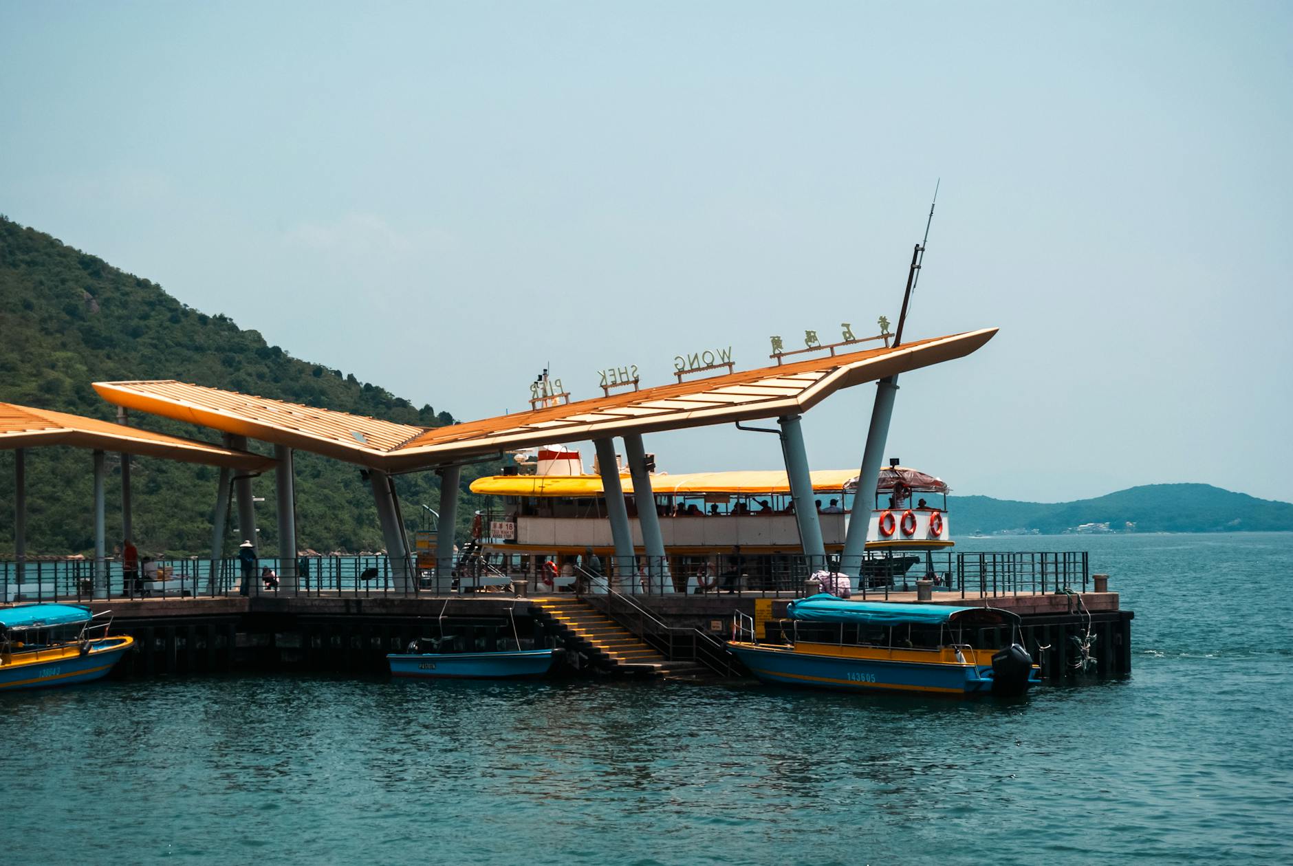Scenic view of a modern pier in Hong Kong with boats, calm water, and a hillside landscape on a sunny day.