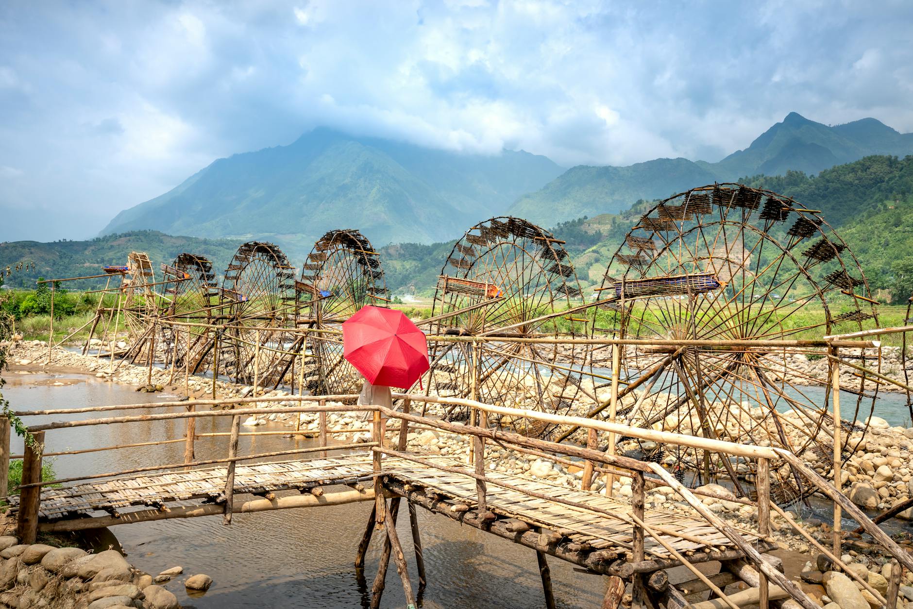 Red umbrella on bamboo fence of footbridge above river with water wheels against misty mountains under cloudy sky