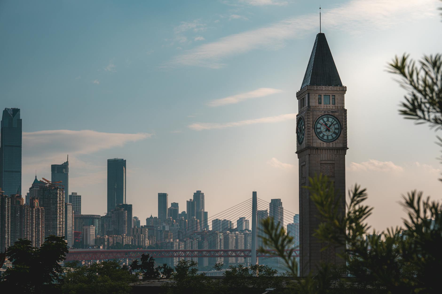 Dramatic city skyline featuring a prominent clock tower at sunset, visible through trees.