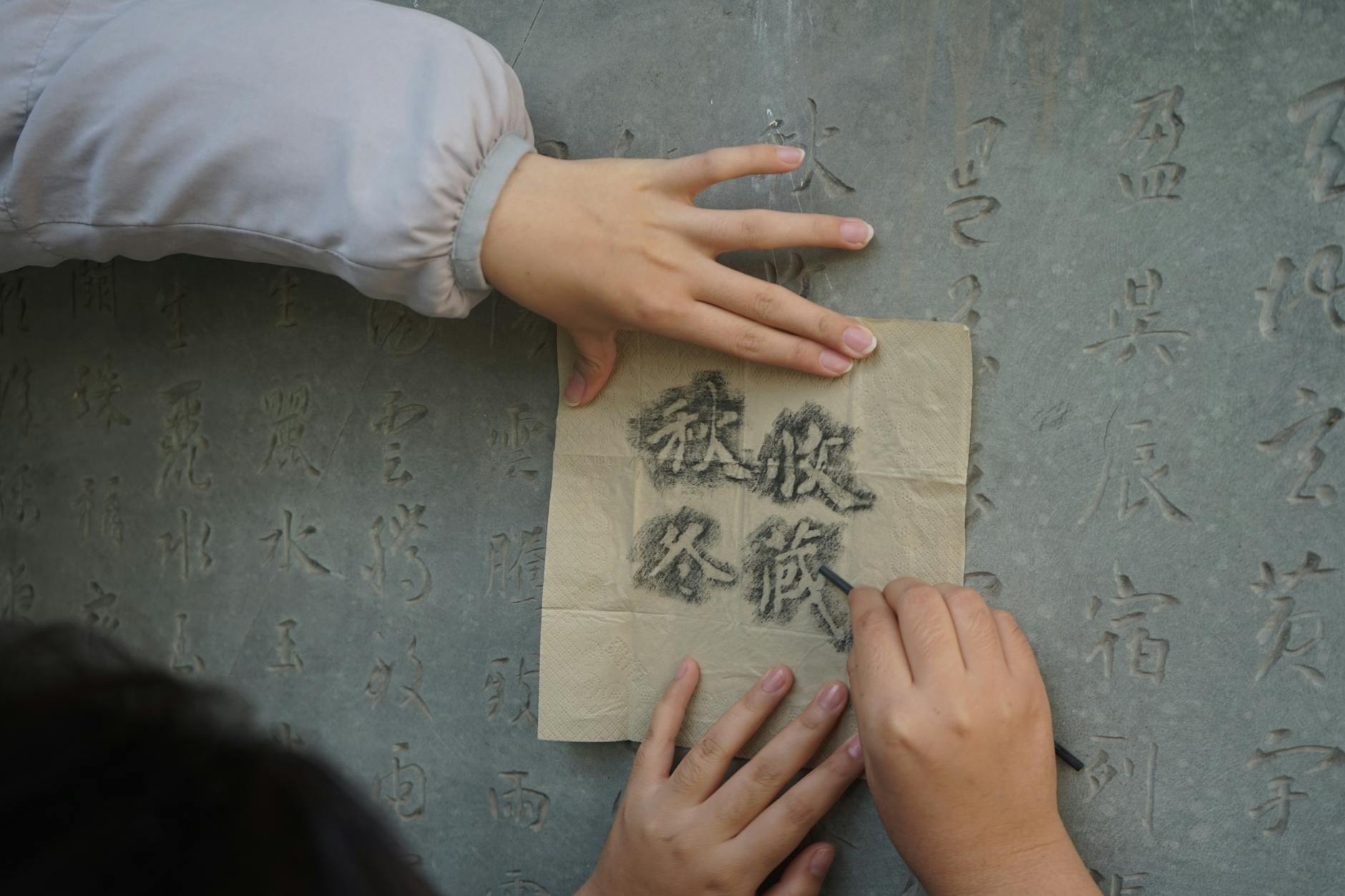 Close-up of hands using charcoal to trace Chinese characters on a stone wall, highlighting calligraphy art.