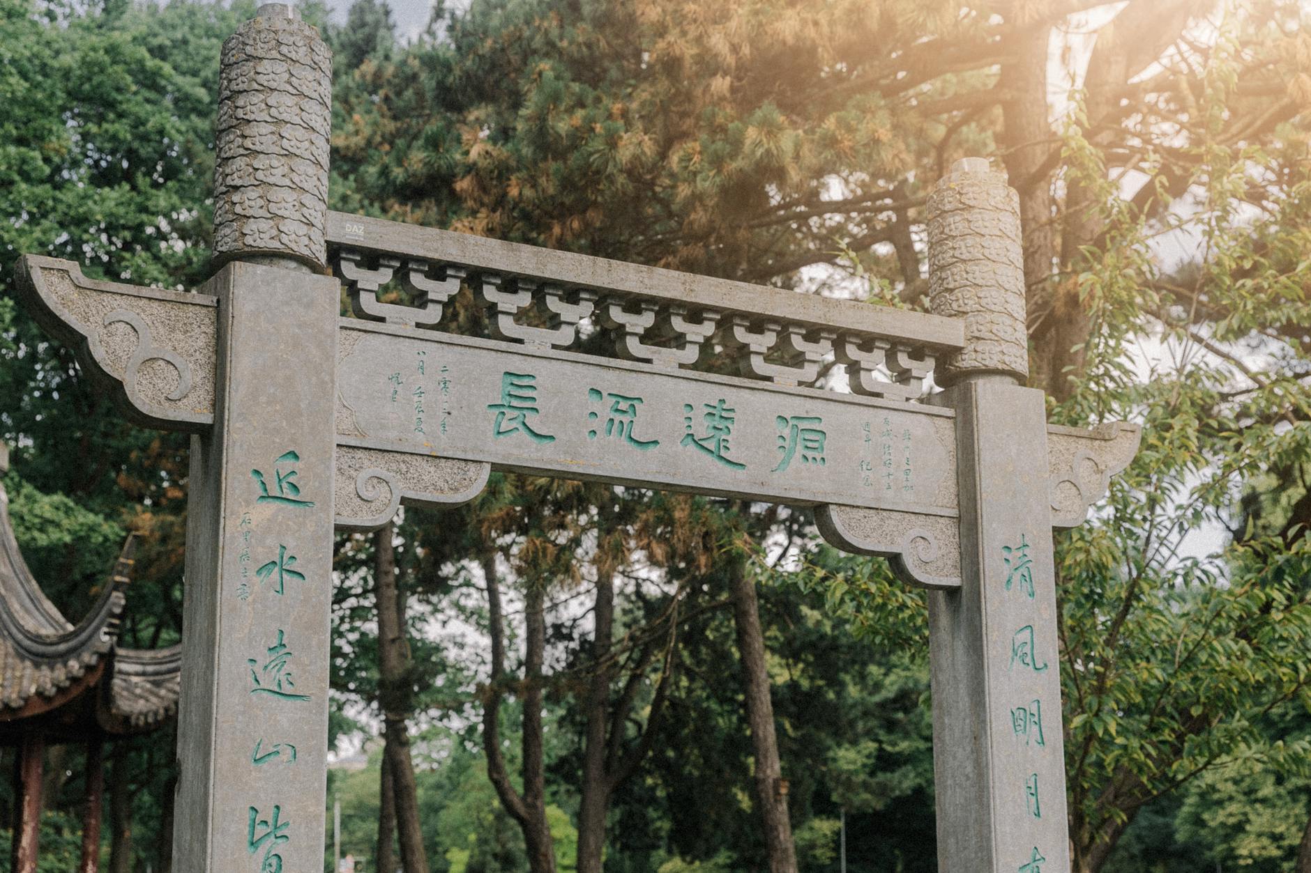 Stone gate with Chinese writings surrounded by lush trees in a serene garden setting.