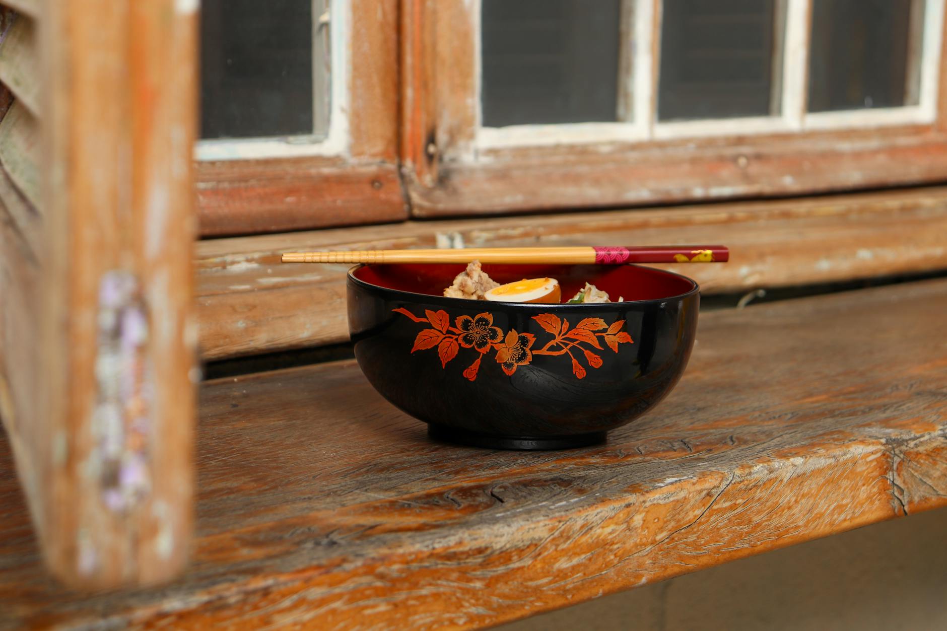 A decorative Japanese bowl with noodles and chopsticks set on a rustic wooden surface indoors.