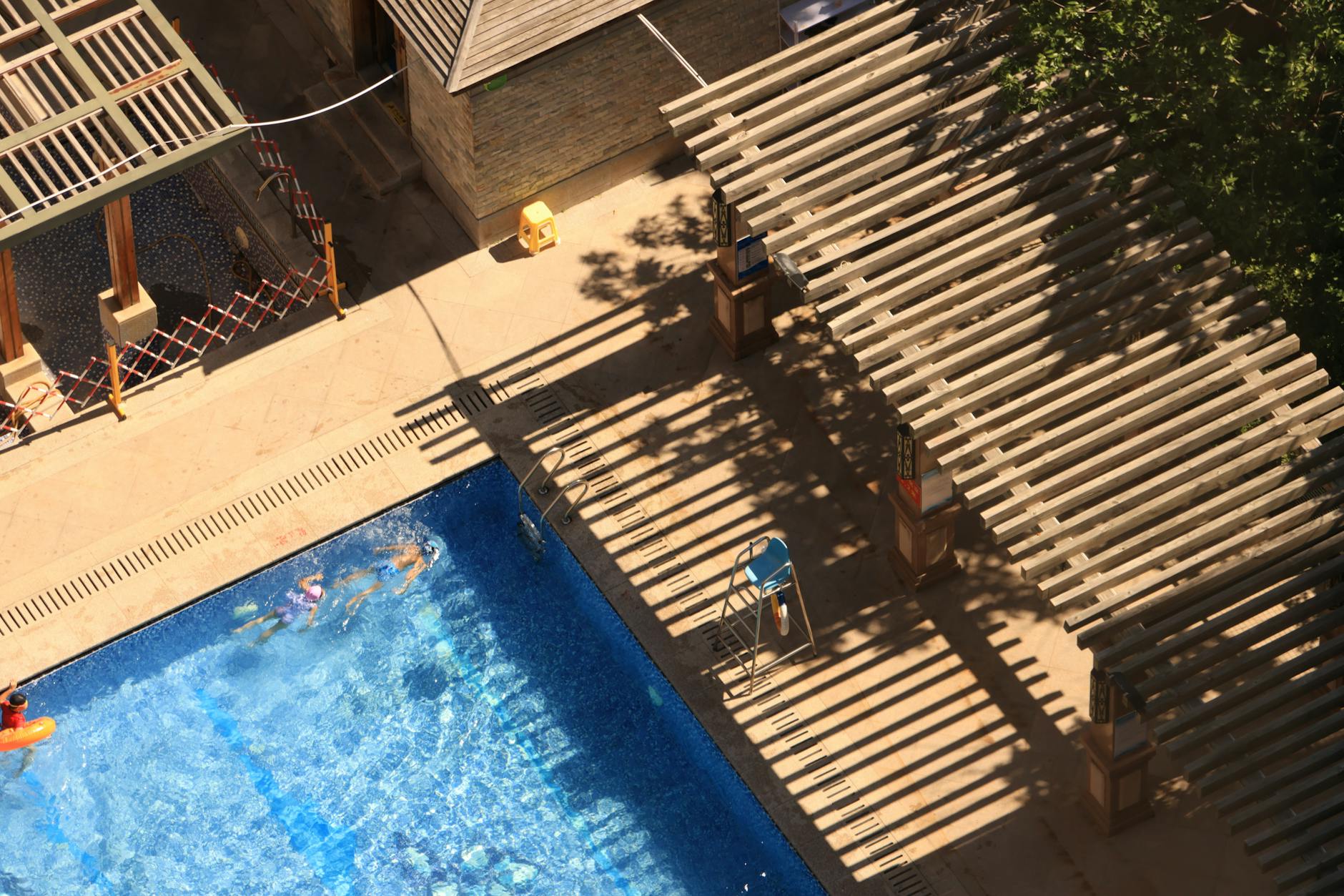 High-angle view of a tranquil resort pool with sunlit pergola and swimmers, perfect for summer vacation vibes.