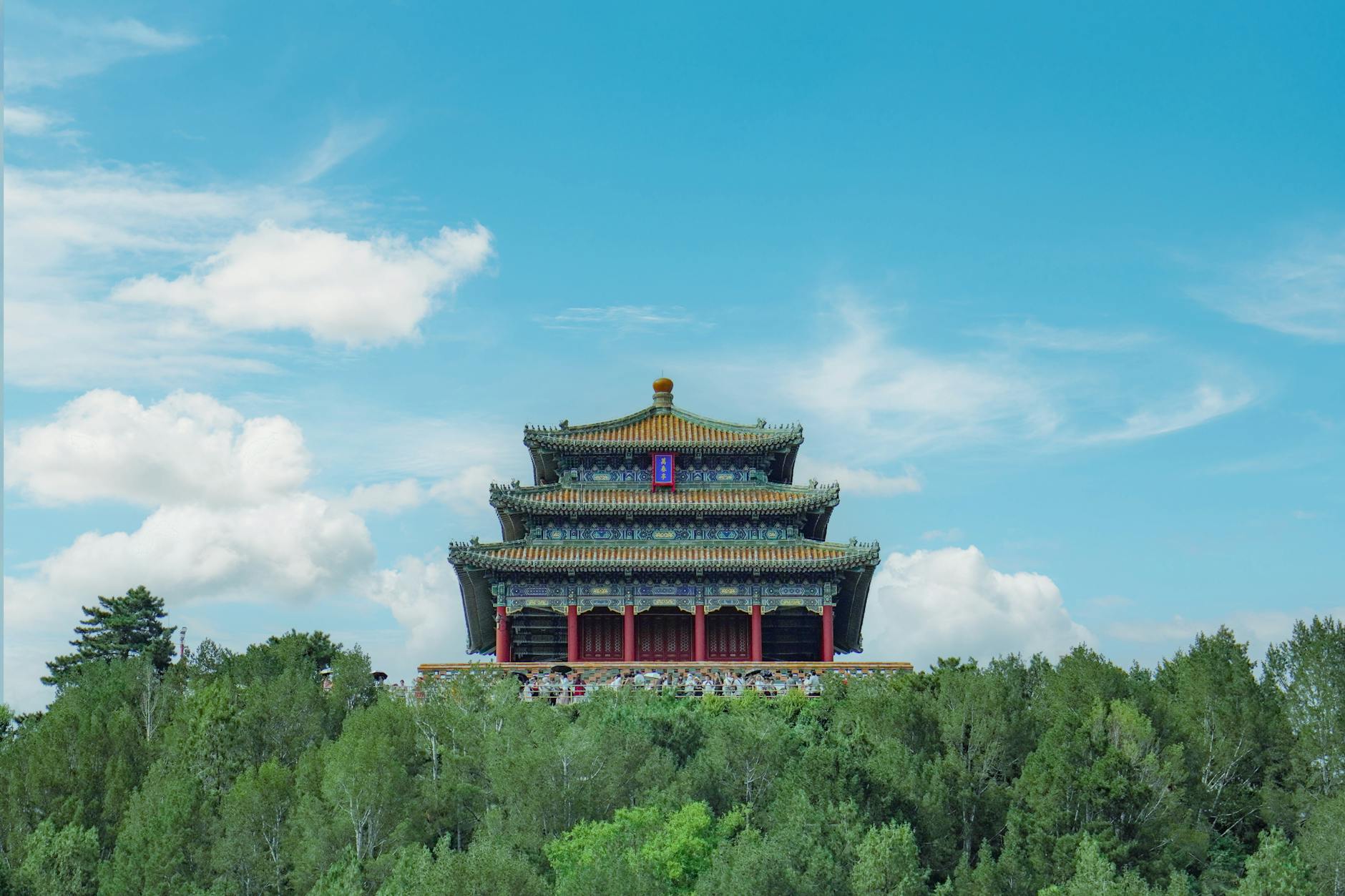 A traditional Chinese pavilion emerges above a lush green forest under a bright blue sky.