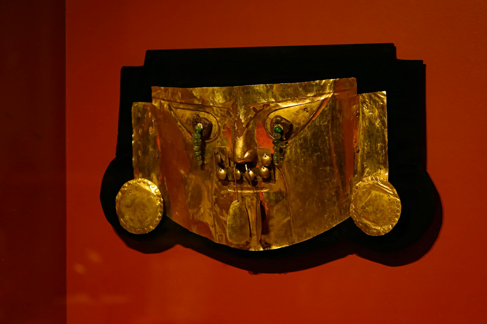 A golden artifact mask displayed on a vibrant red wall, showcasing intricate metalwork.