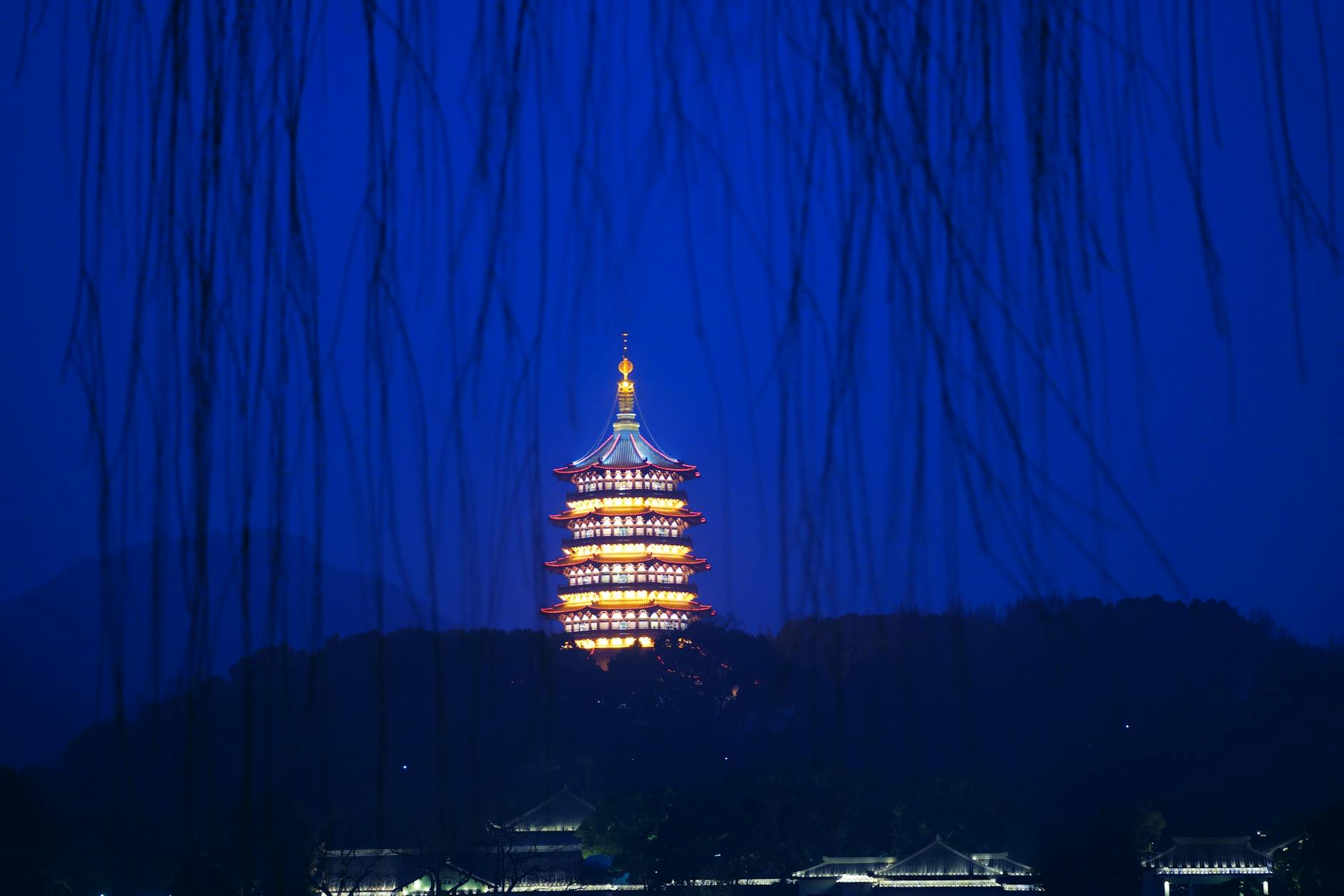 A stunning nighttime view of the illuminated Leifeng Pagoda in Hangzhou, framed by tree branches.