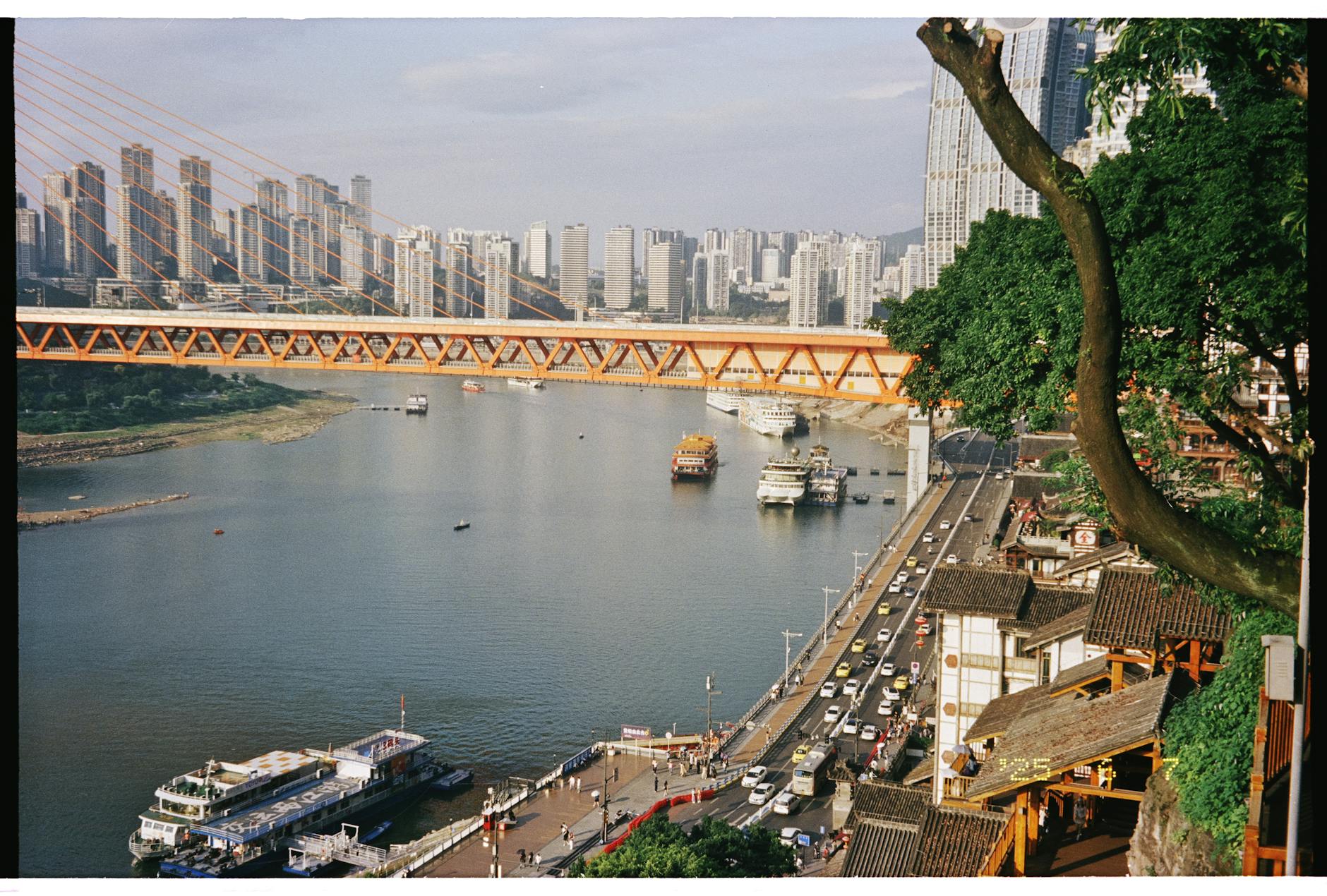 Scenic view of Chongqing city with modern bridge and bustling riverfront activity under the bright daylight.