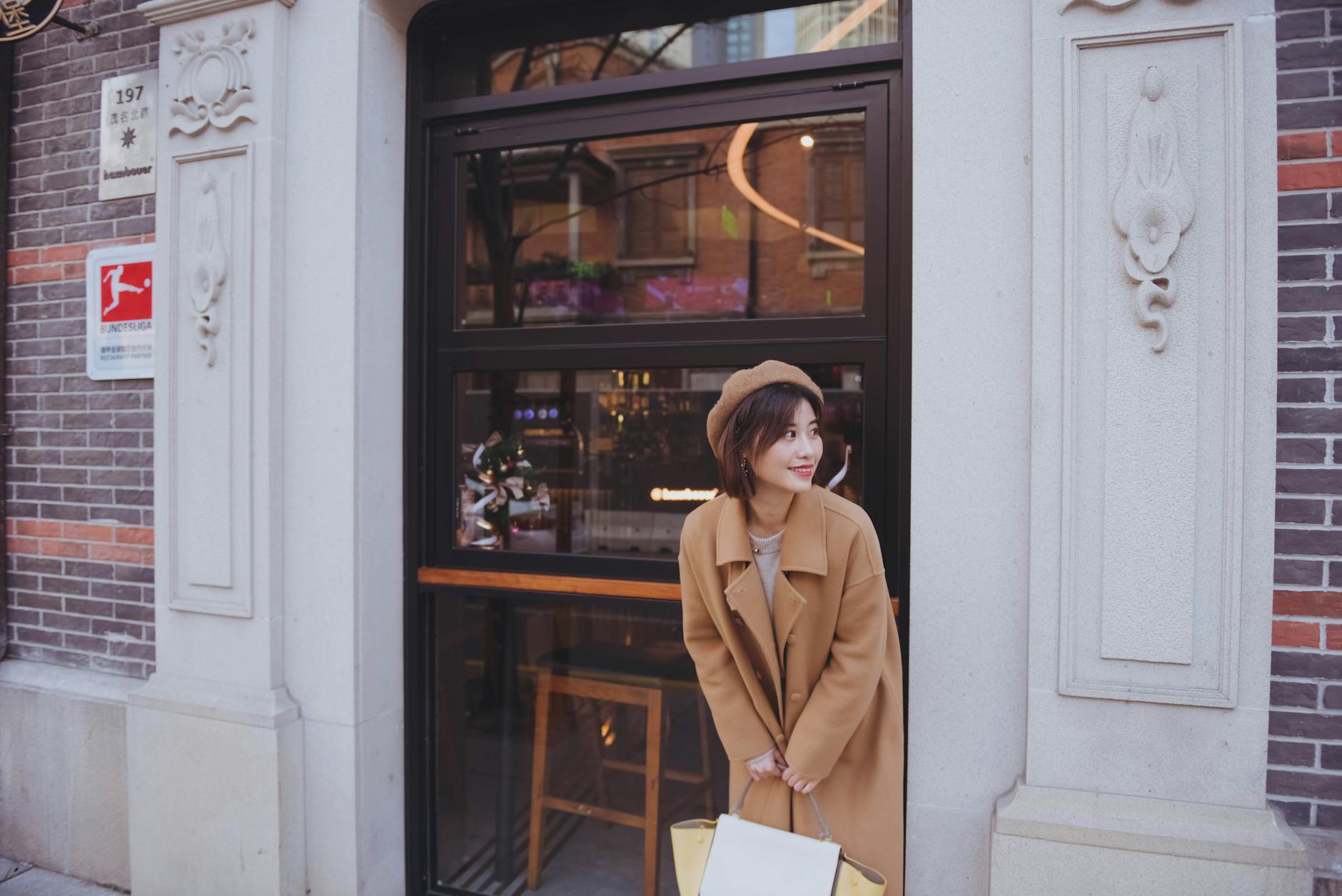 Trendy adult woman in fashionable coat and hat standing outside a modern Shanghai café entrance.