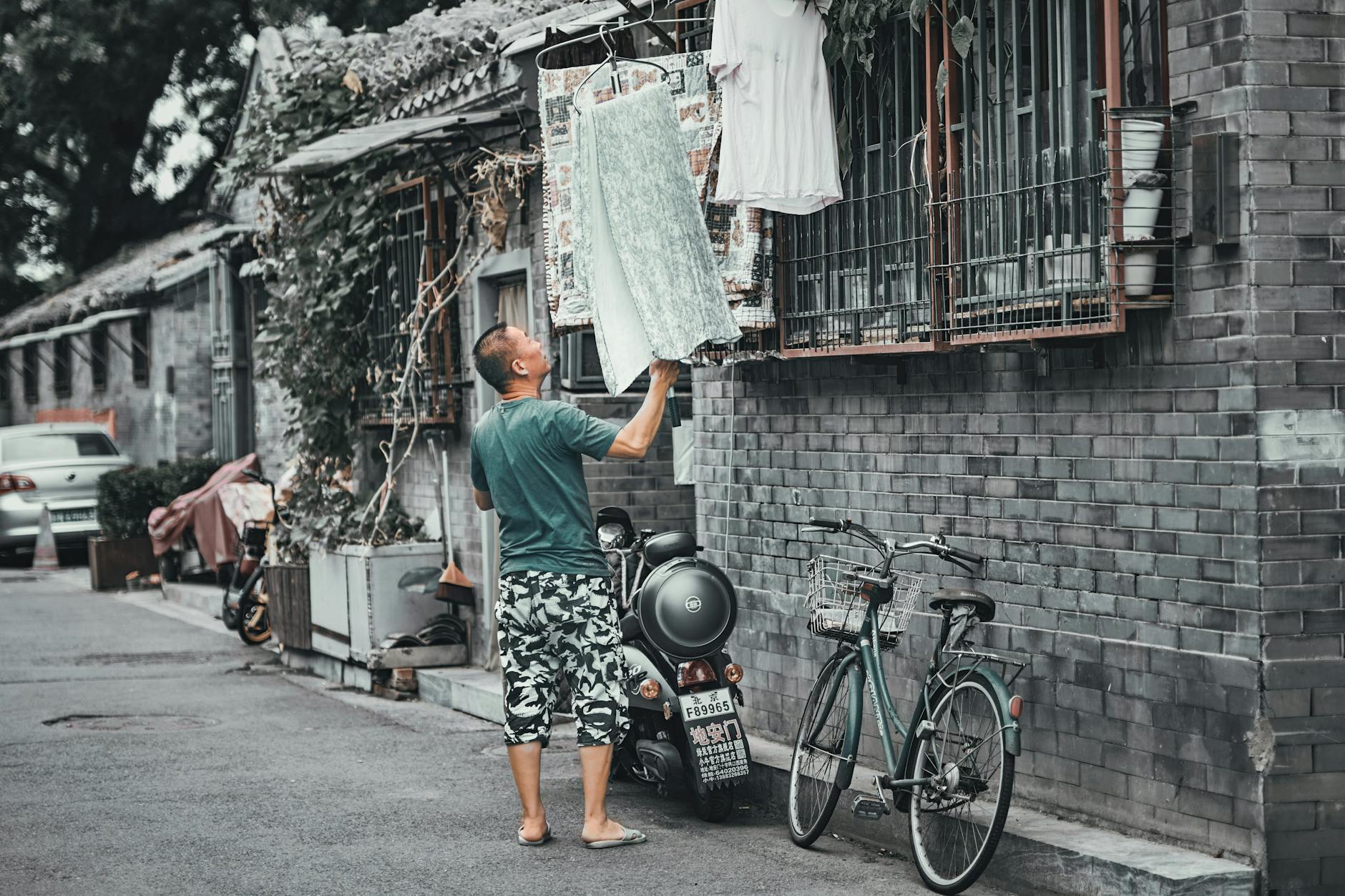 A man hangs laundry outside his house in a traditional Beijing hutong, with a bicycle and motorcycle nearby.