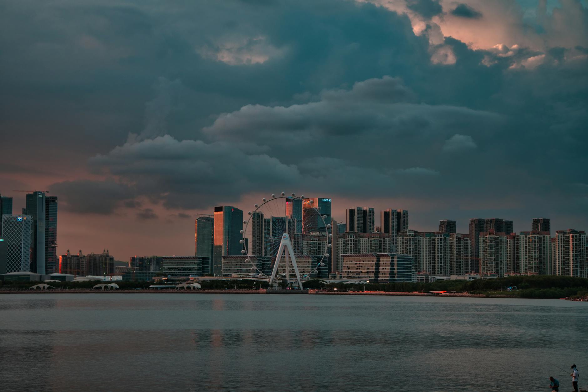 Breathtaking view of Shenzhen's waterfront cityscape with Ferris wheel, captured at twilight under dramatic clouds.
