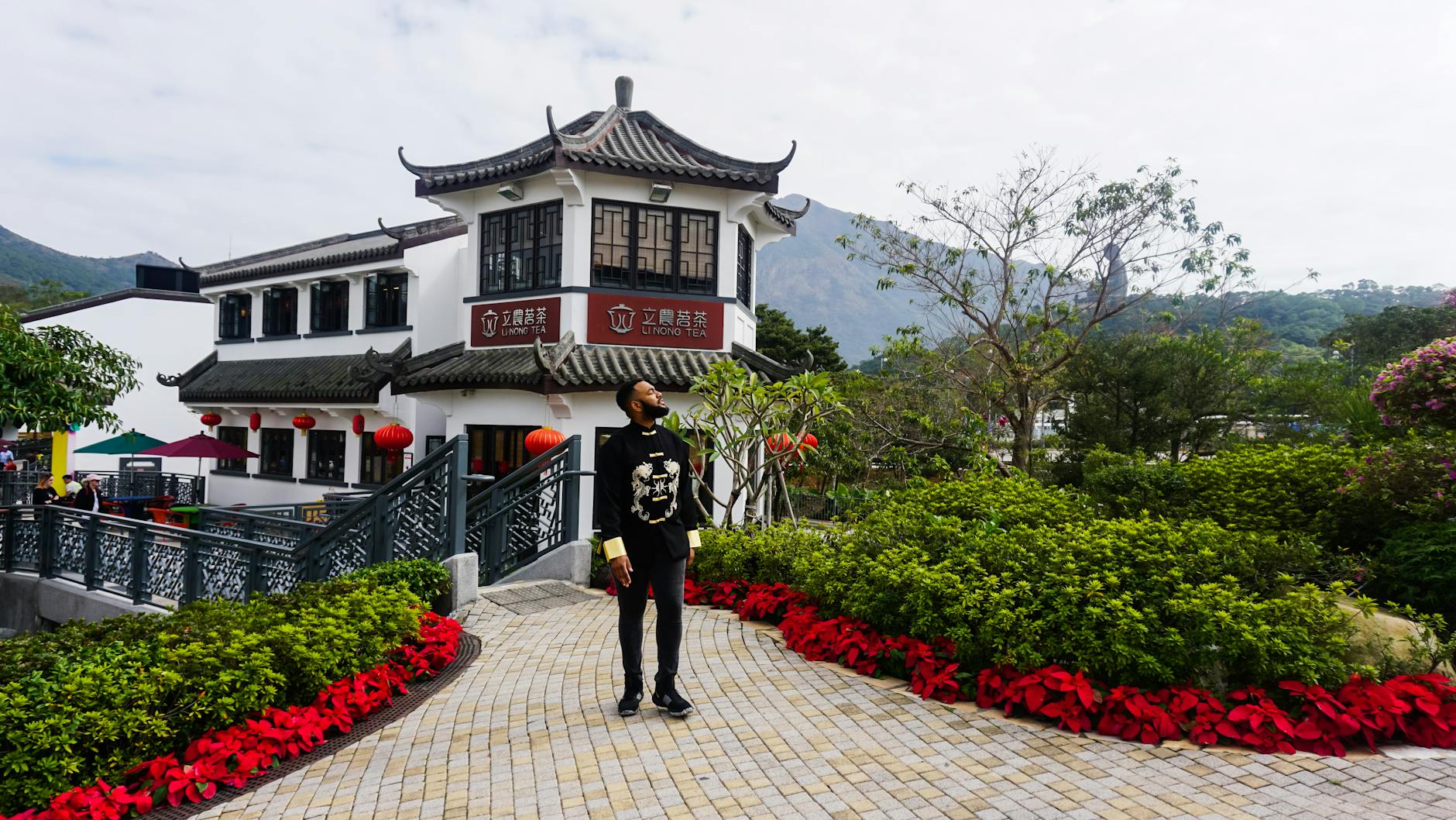A man stands on a walkway in Ngong Ping Village, Hong Kong, with traditional architecture and lush gardens.