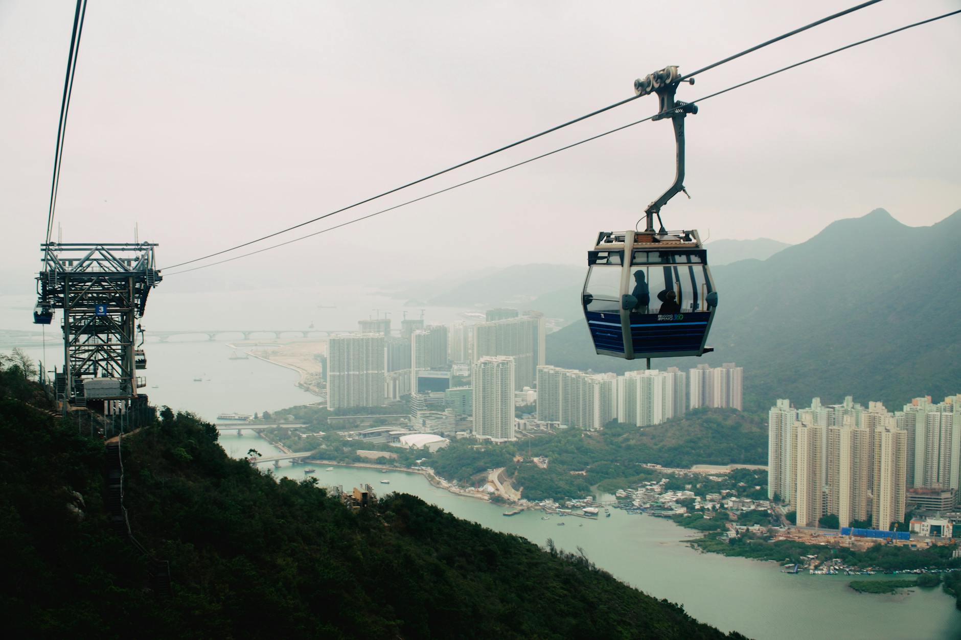 Cable car travels over lush hills with a cityscape view, offering a blend of urban and natural beauty.