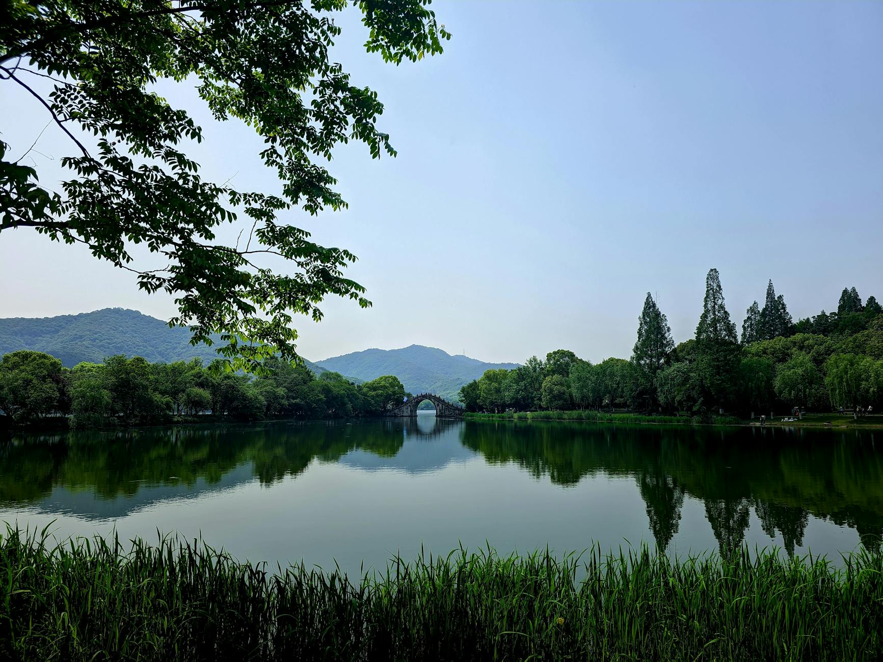 A peaceful lake scene featuring an arch bridge and lush greenery under a clear sky.