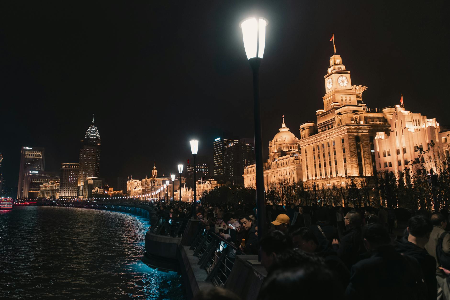 Low angle of aged masonry Bund exterior with clock and national flag near river and glowing lamp post above unrecognizable people in town at dusk