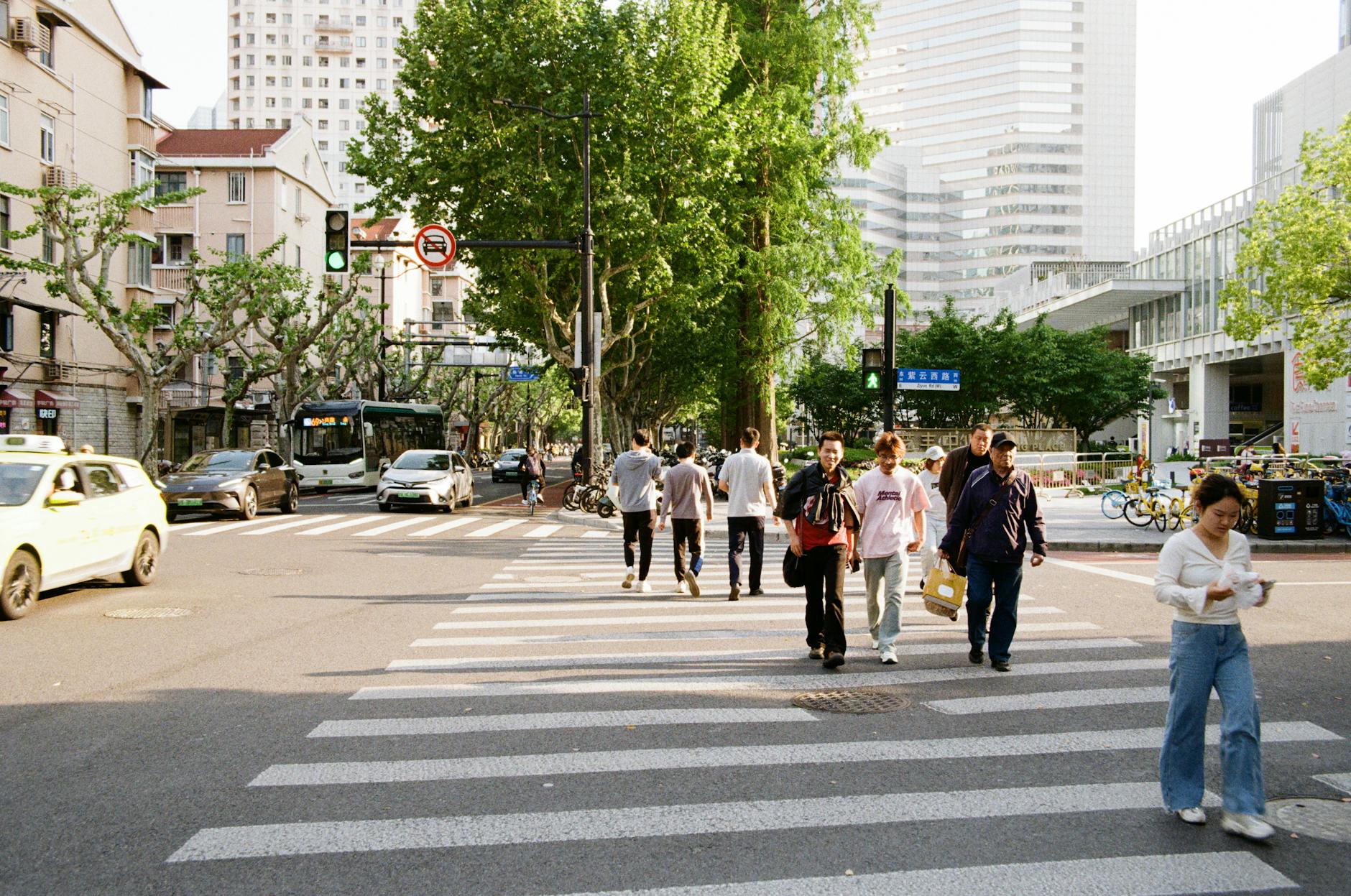 Pedestrians crossing a bustling street in Shanghai with urban buildings and greenery in the background.