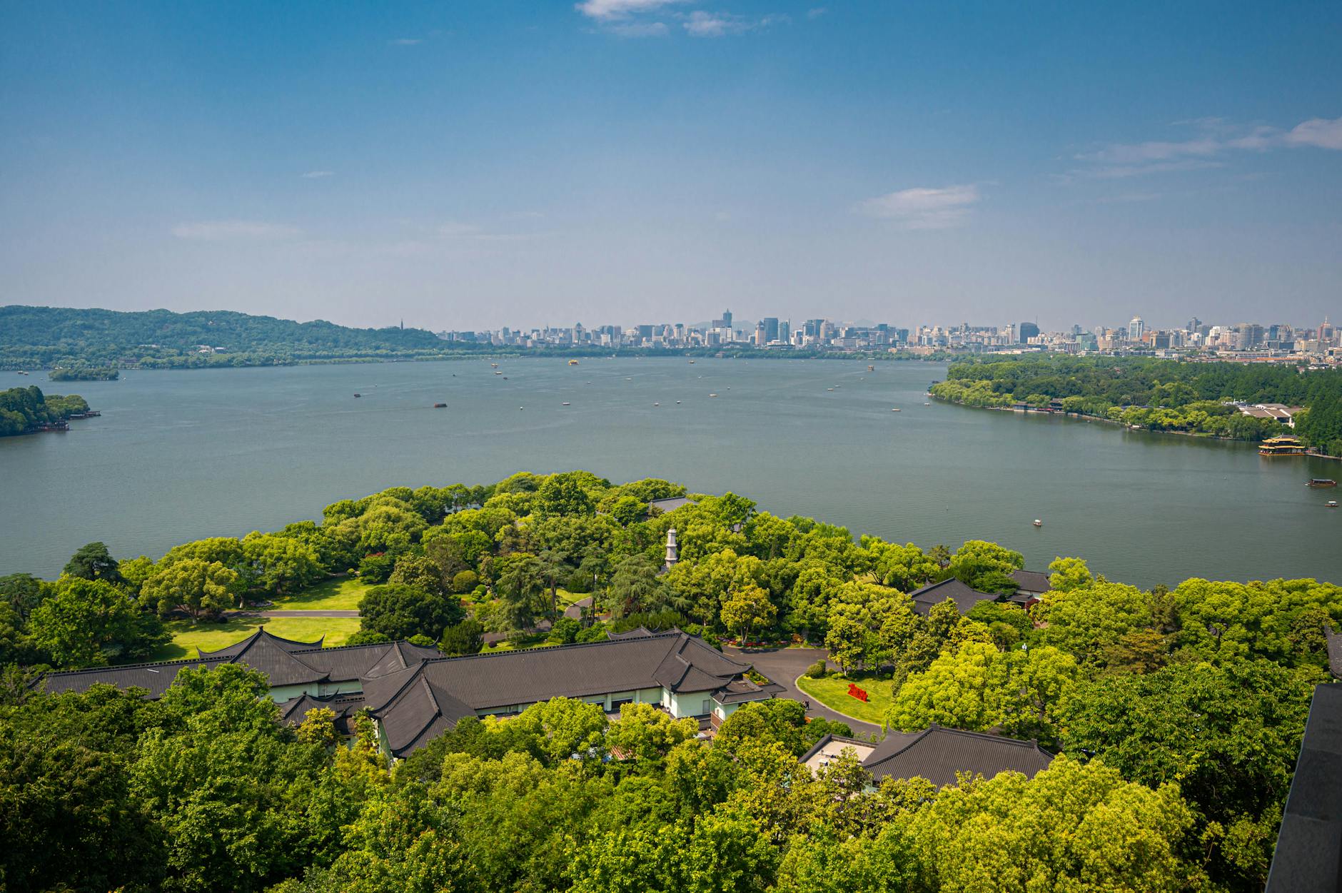 Aerial view over West Lake, lush greenery, and Hangzhou city skyline under a clear blue sky.