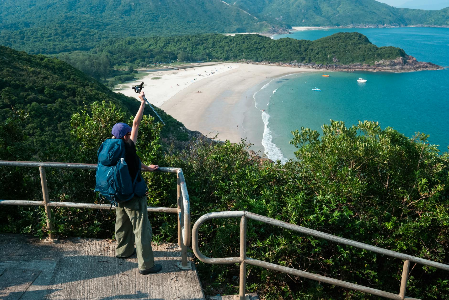A hiker with a backpack photographs a breathtaking coastal view from a railinged viewpoint.