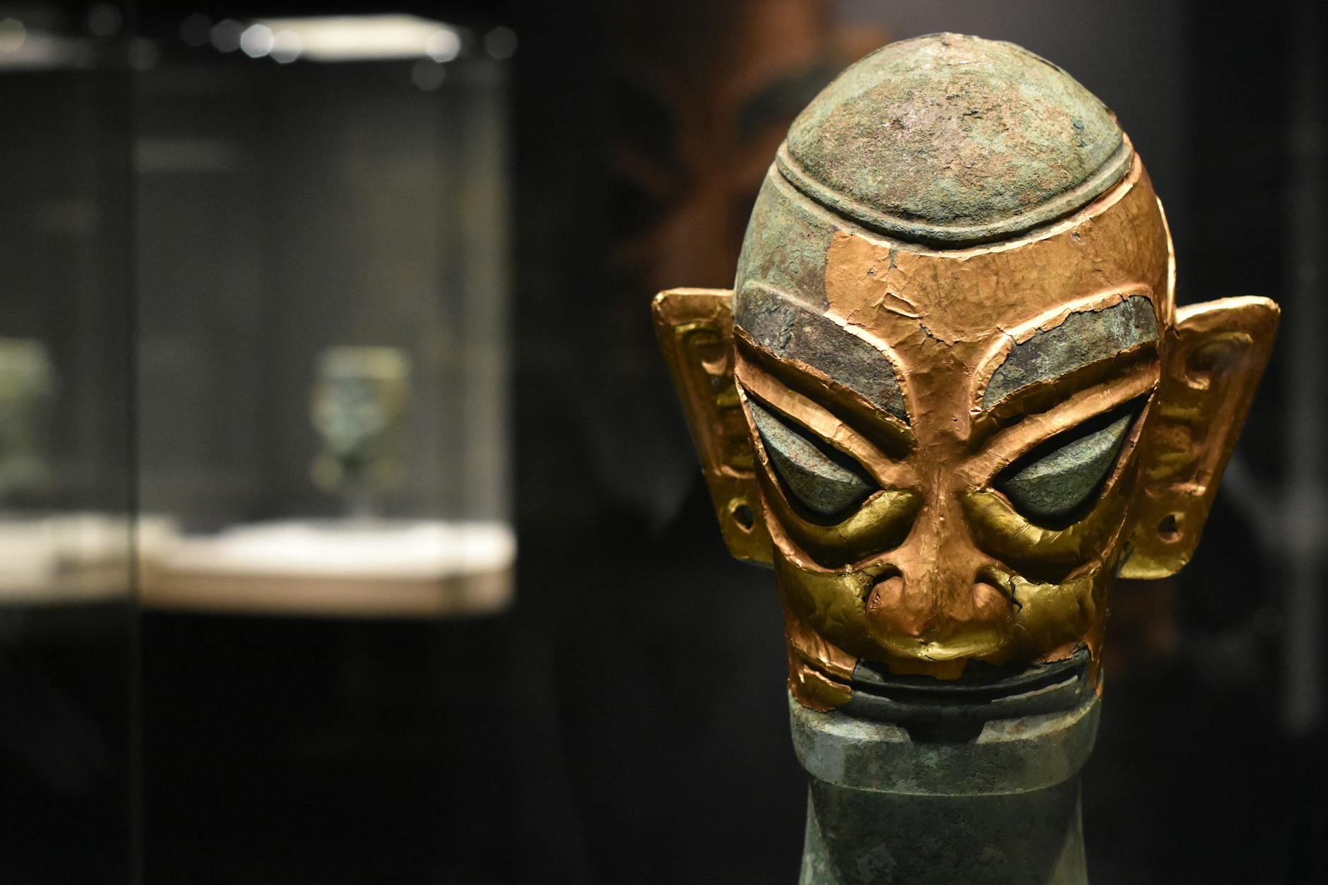 Close-up of an ancient bronze mask exhibited in a dimly lit museum display case.