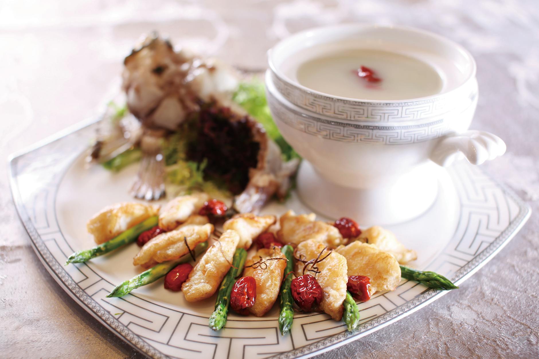 Elegant seafood dish featuring asparagus, fried fish, and a creamy soup presented on a decorative plate.