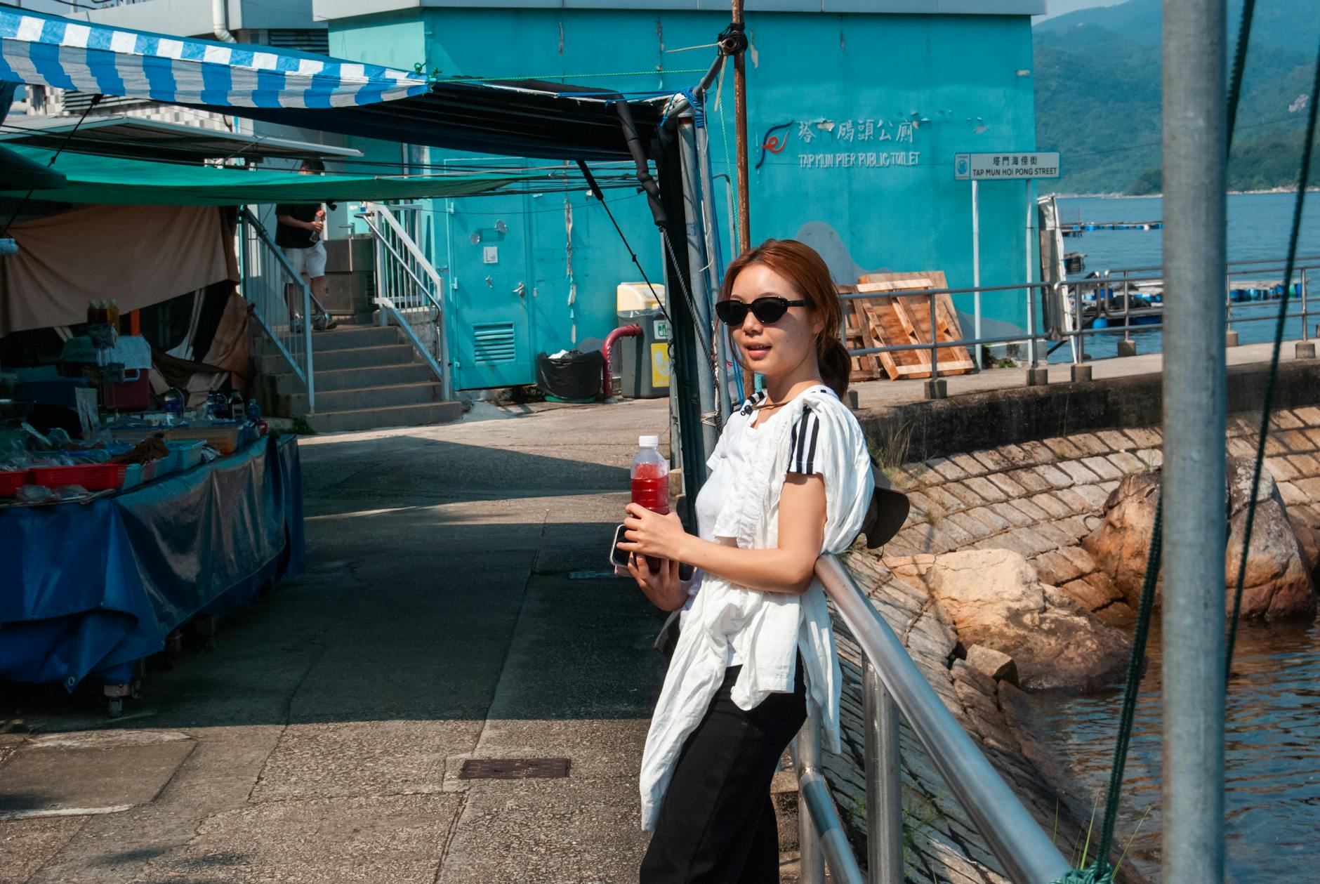 Woman in sunglasses at Hong Kong waterfront, enjoying a sunny day with a drink. Vibrant and casual outdoor scene.