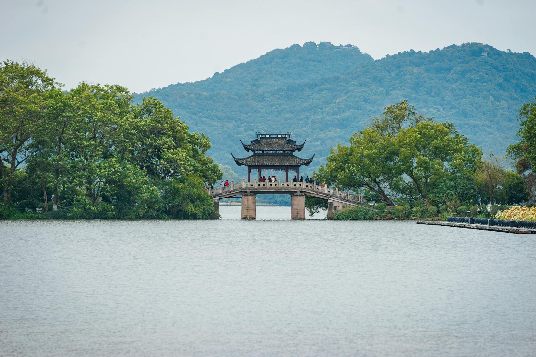 A serene view of a traditional pavilion on a bridge surrounded by lush greenery and mountains in Hangzhou, China.
