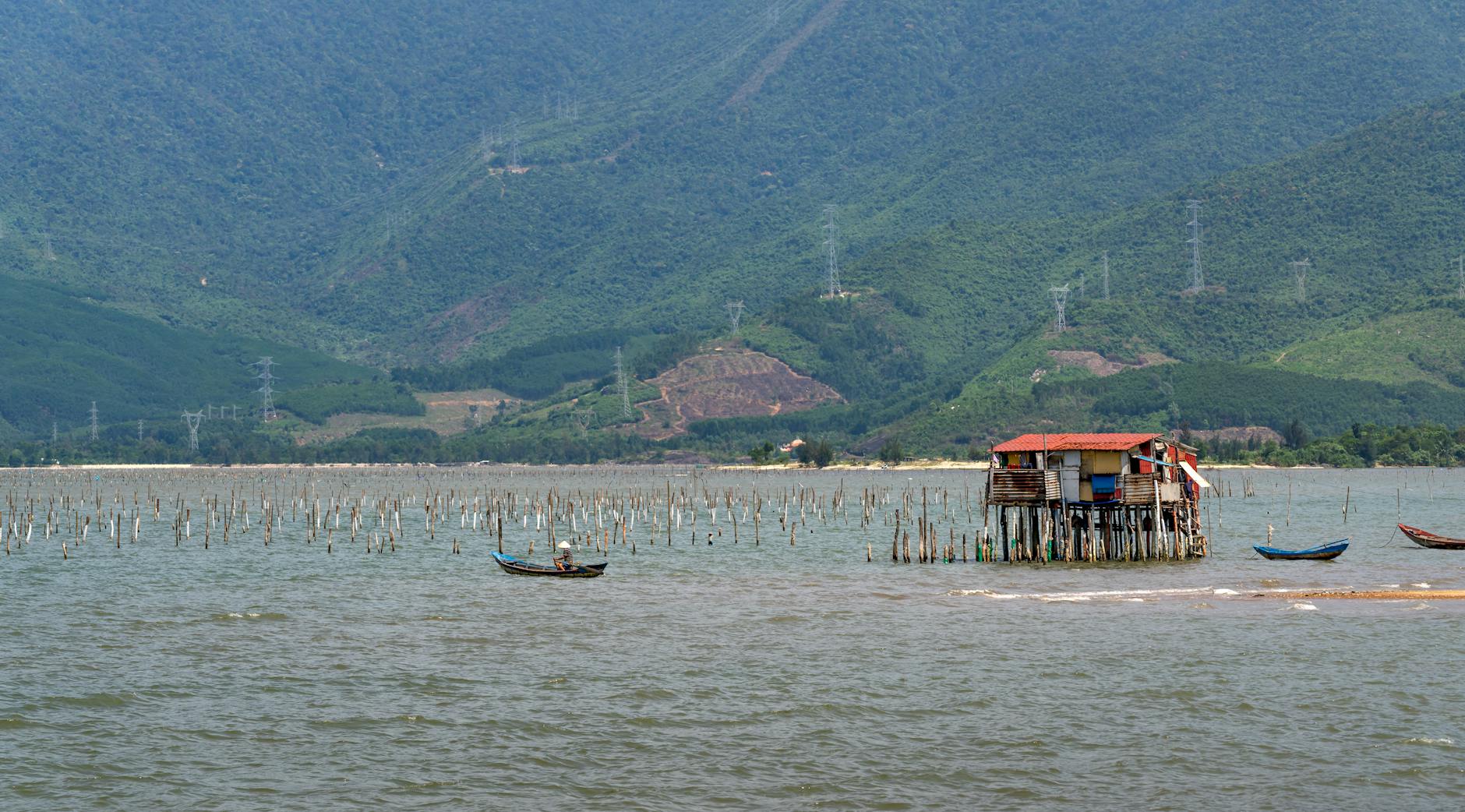 Scenic view of a stilt house in a lagoon with a boat and lush green hills in the background.
