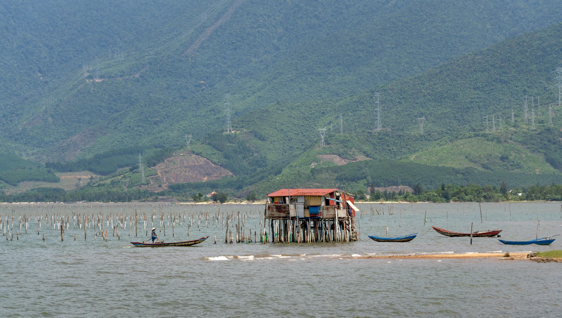 Fisherman in traditional boat near stilt house against mountain backdrop.