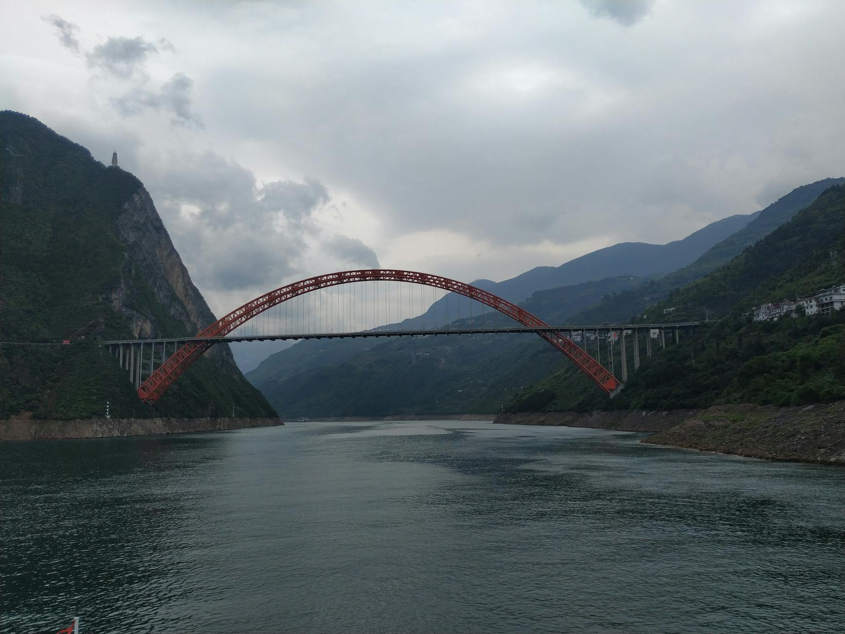 Stunning red arch bridge over a wide river with lush mountains beneath a cloudy sky.