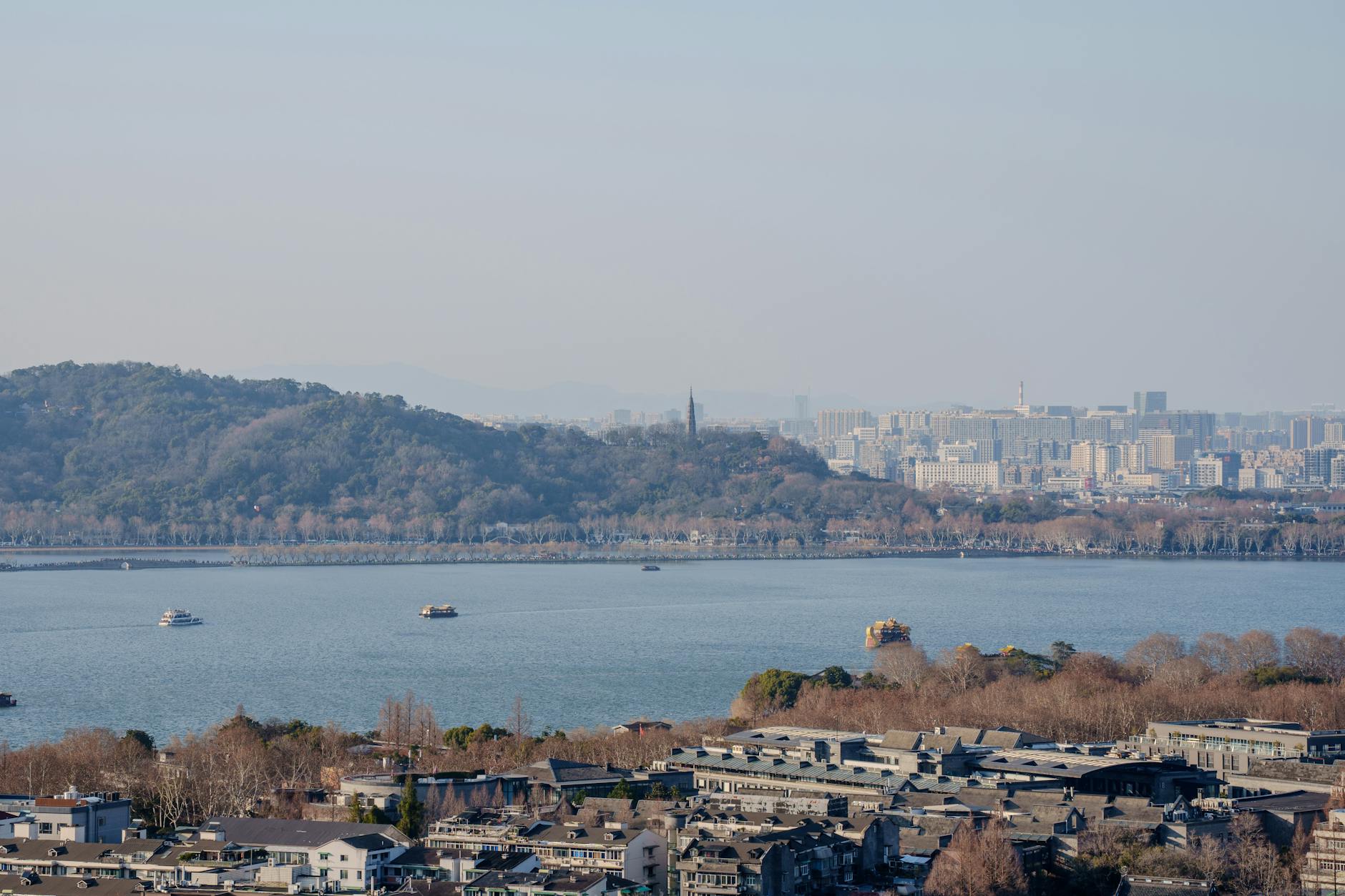 Aerial view of West Lake, Hangzhou with distant city skyline and boats on a sunny day.