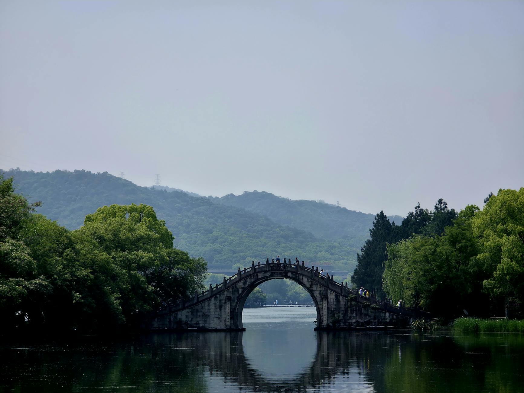 Serene landscape featuring a traditional stone arch bridge over a calm lake, with lush greenery and distant mountains.