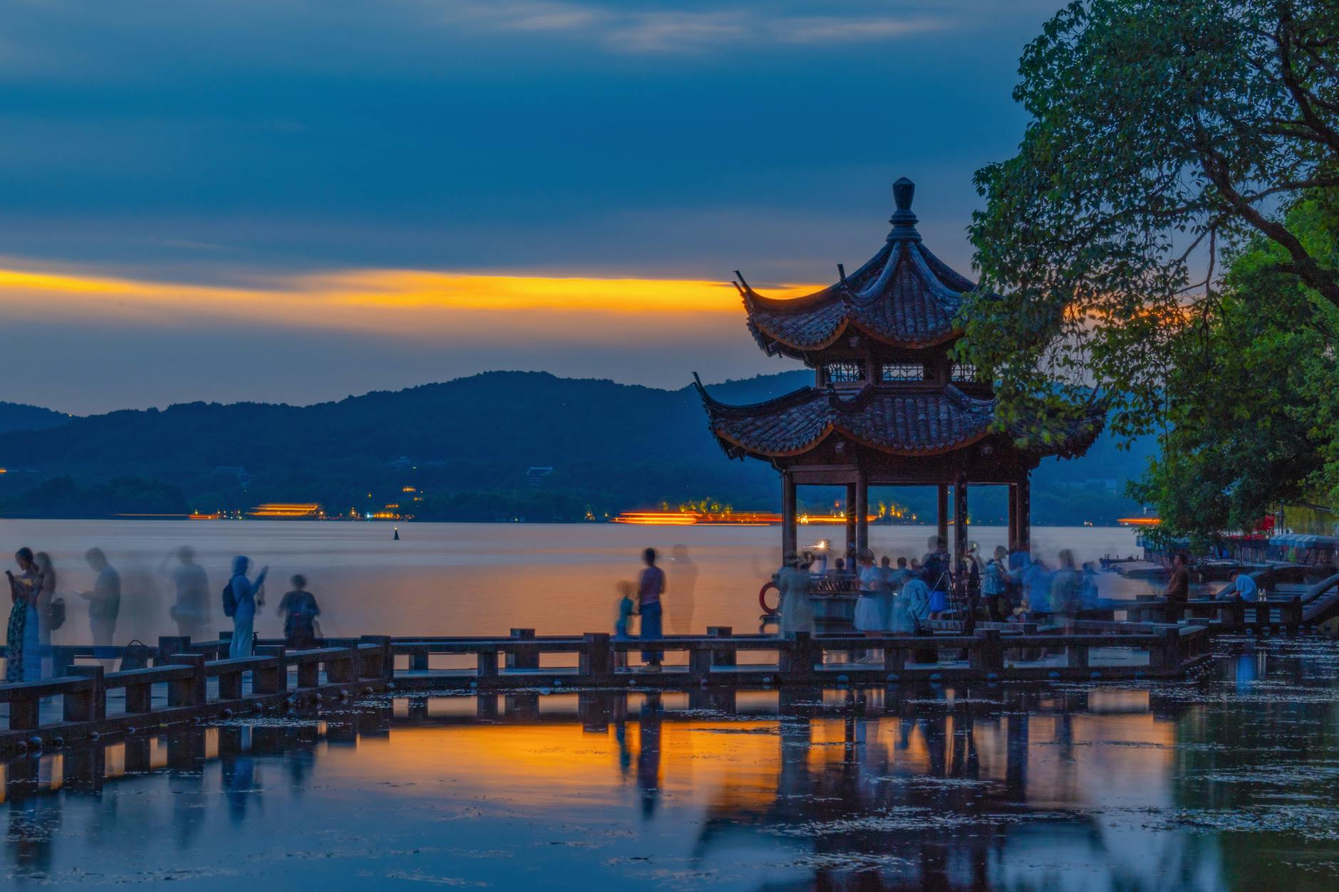 Serene sunset at West Lake with a traditional Chinese pavilion and blurred people.