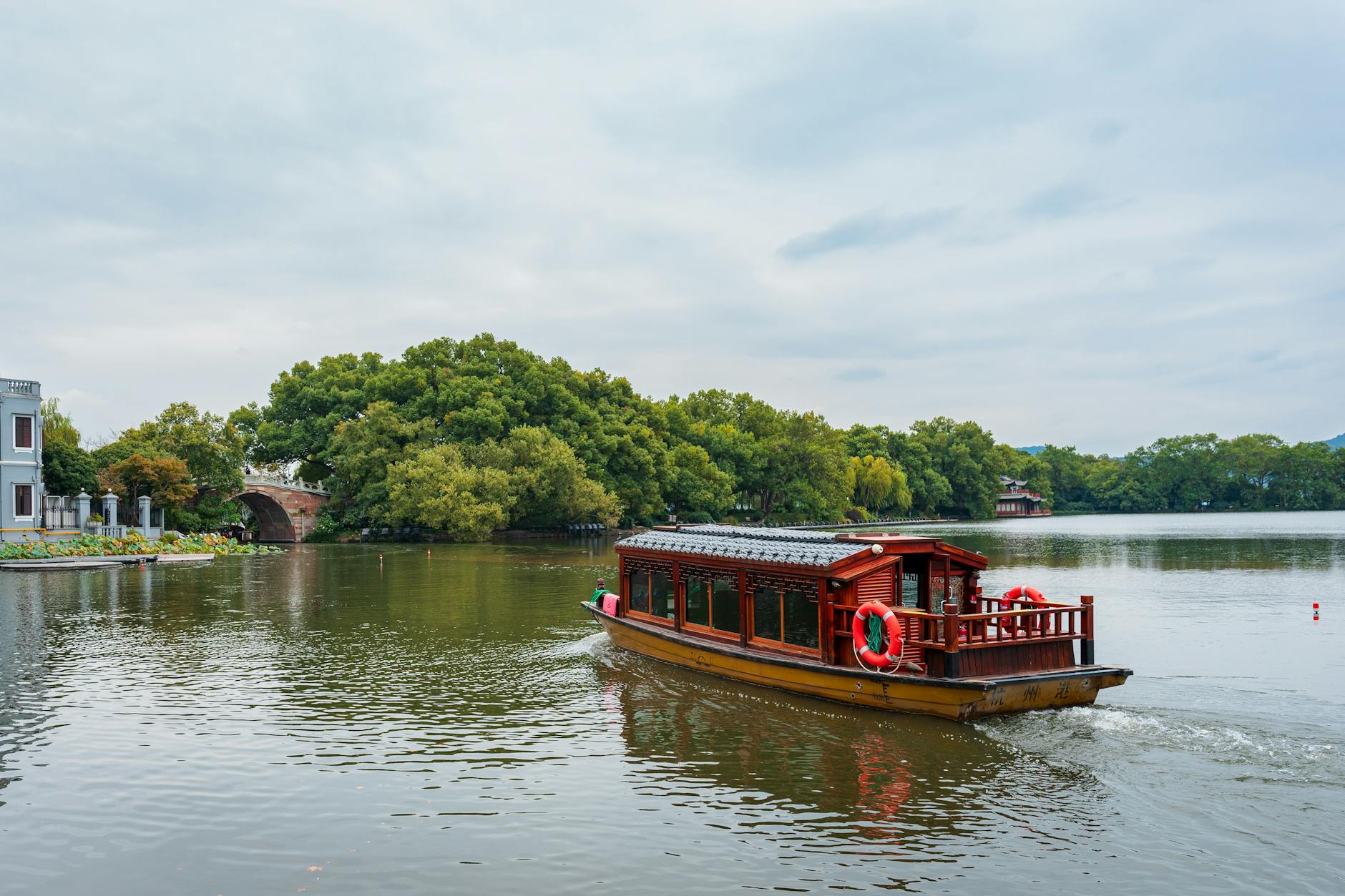 Scenic view of a traditional wooden boat on West Lake, Hangzhou, highlighting lush greenery and calm water.