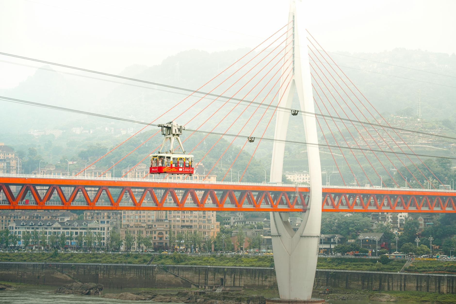 A cable car crosses the Yangtze River bridge in Chongqing, featuring vibrant architecture and lush hills in the background.